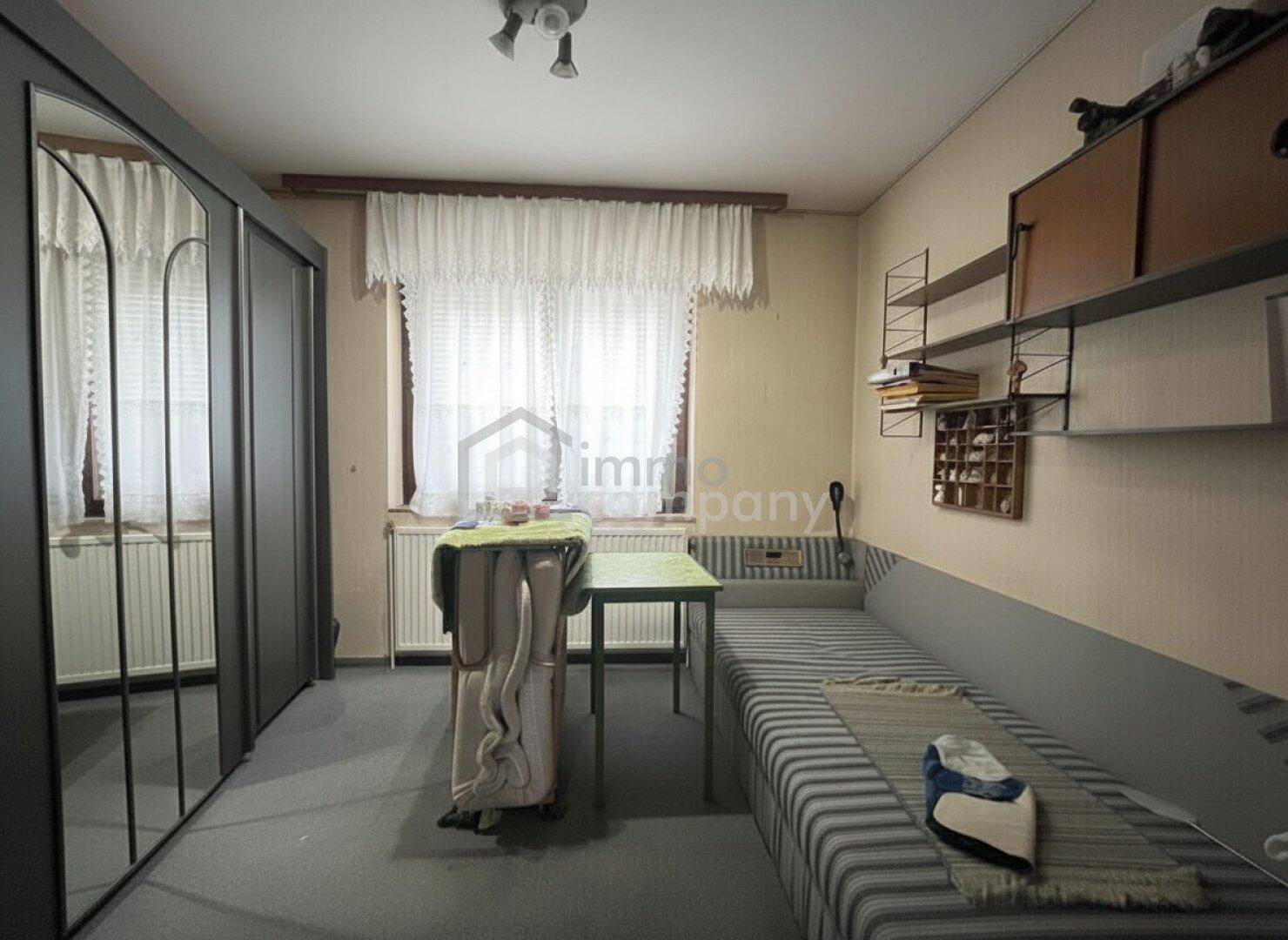 Einfaches Schlafzimmer mit Einzelbett, Kleiderschrank und Fenster mit Vorhängen.