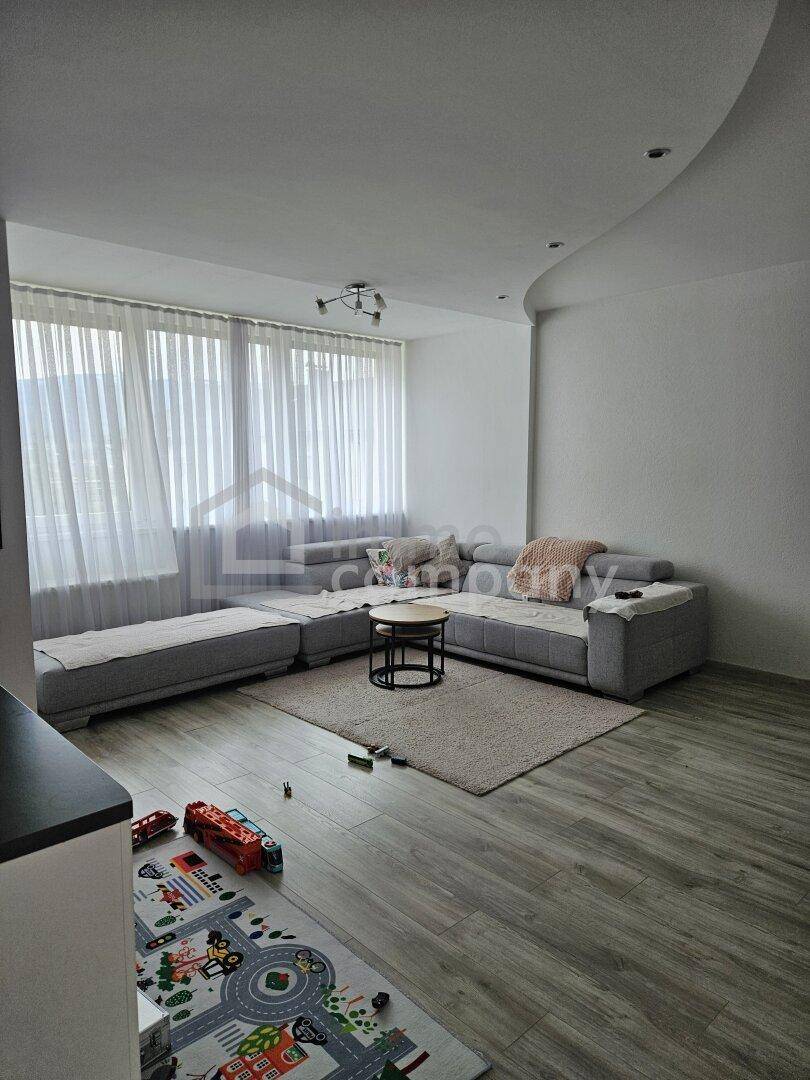 Gemütliches Wohnzimmer mit großem Ecksofa, Teppich und breiten Fenstern mit Vorhängen.