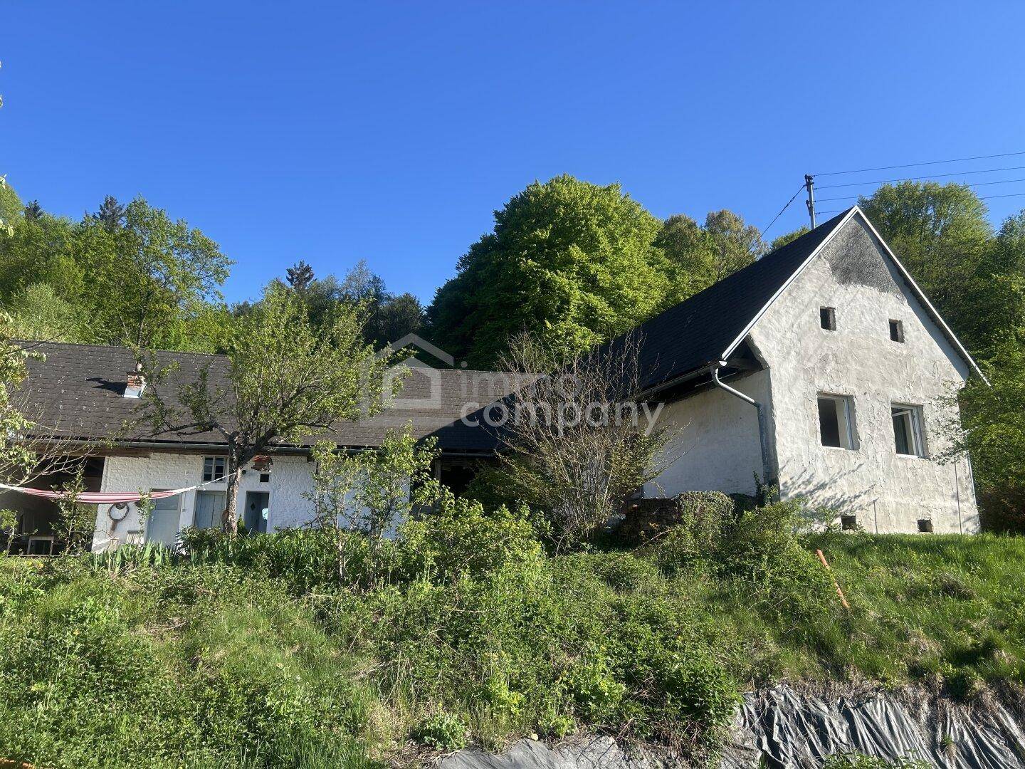 Ansicht eines ländlichen Gebäudes mit dunklem Dach und weißer Fassade, eingebettet in eine grüne Hügellandschaft.