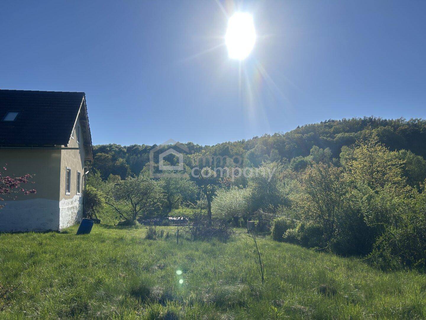 Ländliches Gebäude mit sonniger Wiese und bewaldeten Hügeln im Hintergrund unter einem strahlend blauen Himmel.