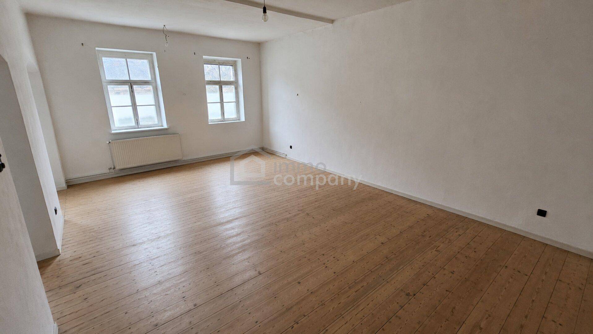 Leerer Raum mit Holzdielenboden, weißen Wänden und zwei Fenstern.