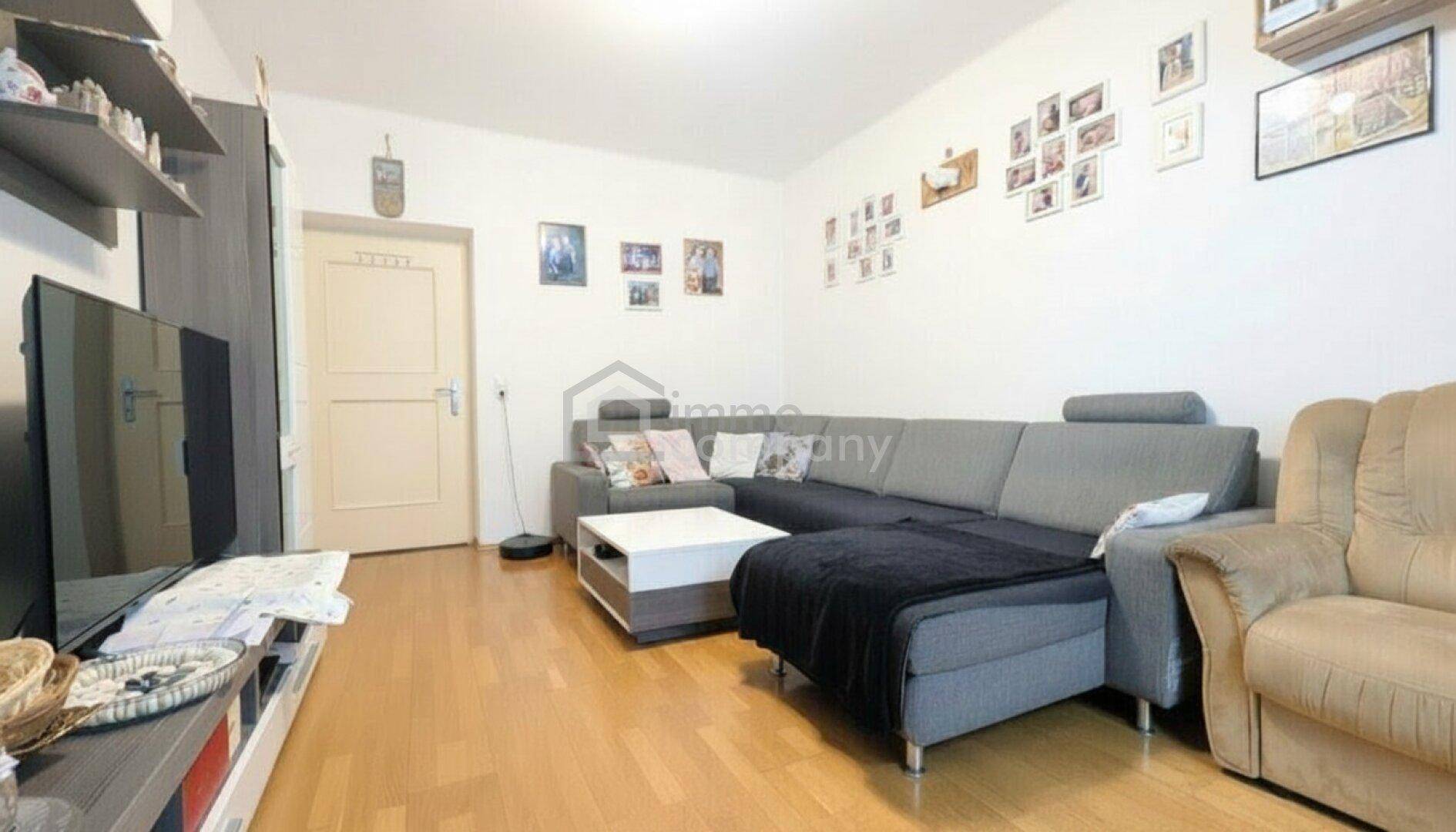 Helles Wohnzimmer mit bequemer Couch, Holzboden und einer Tür, die zu einem anderen Raum führt.