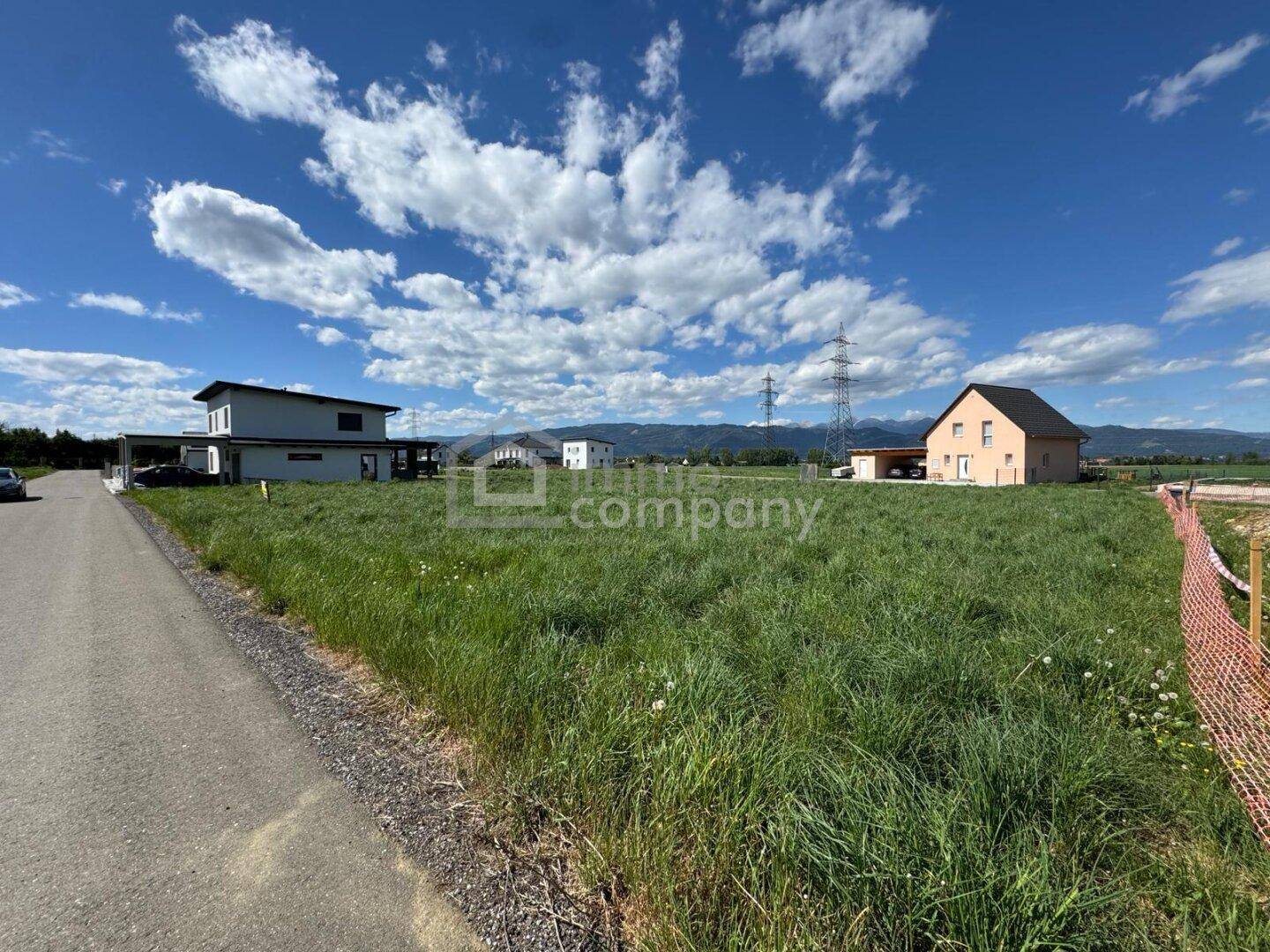Weitläufiges Grundstück mit Grasfläche, umgeben von Wohnhäusern und einer Bergkulisse unter blauem Himmel.