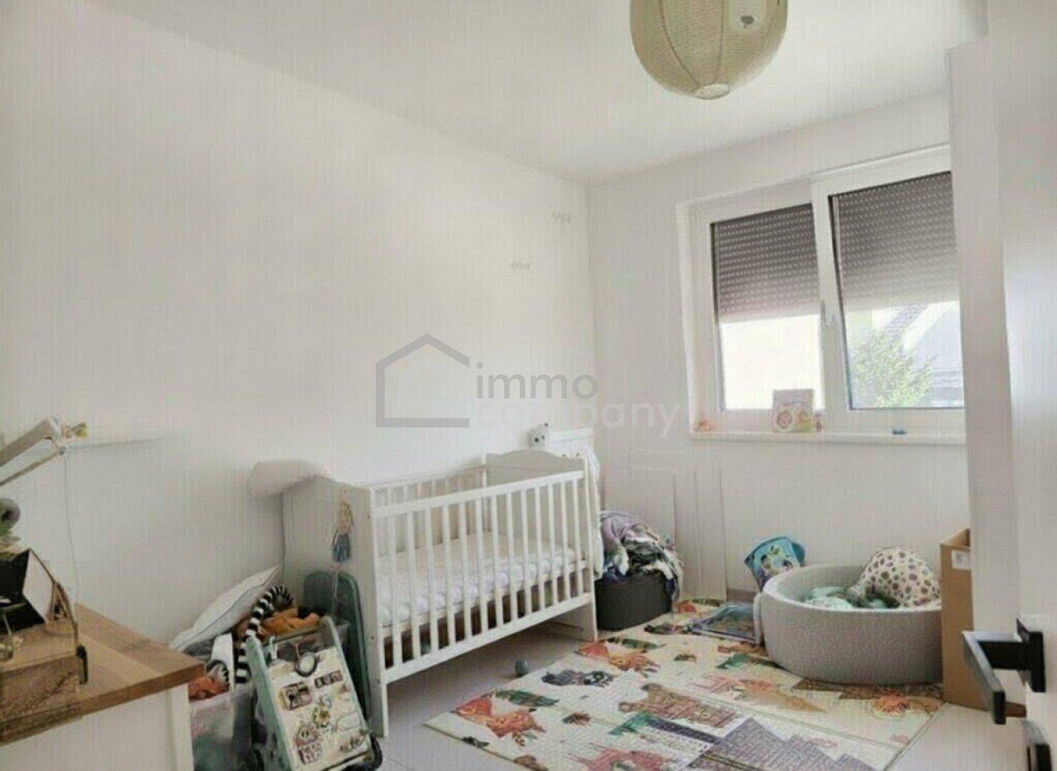 Helles Kinderzimmer mit Babybett, farbenfrohem Teppich und Fenster mit Rollladen.