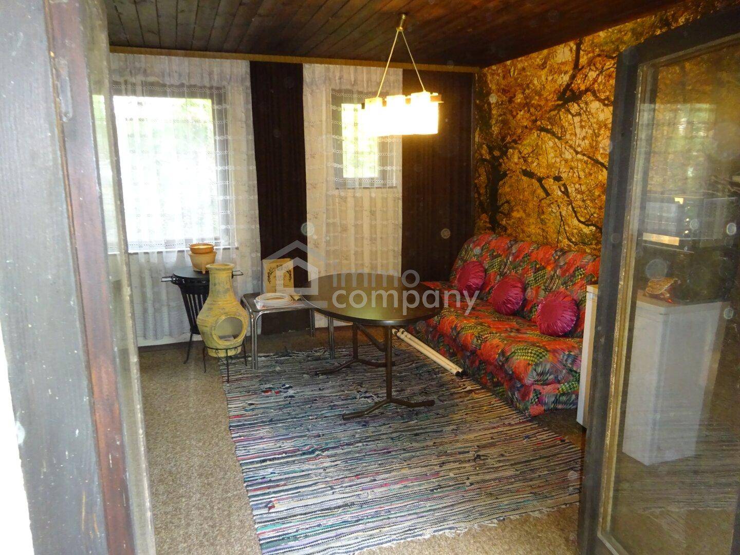 Gemütliches Wohnzimmer mit floralem Sofa, Holzbalkendecke und Teppichboden, durch Fenster hell beleuchtet.