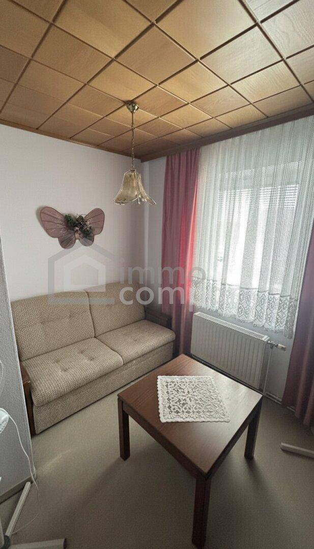 Wohnzimmer mit beigem Sofa, Holztisch und Fenster mit Vorhängen, unter einer dekorativen Holzdecke.