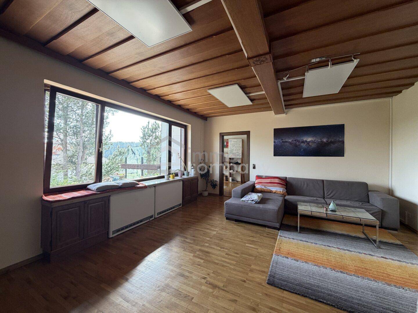 Helles Wohnzimmer mit Holzbalkendecke, Parkettboden und großen Fenstern mit Blick ins Grüne.