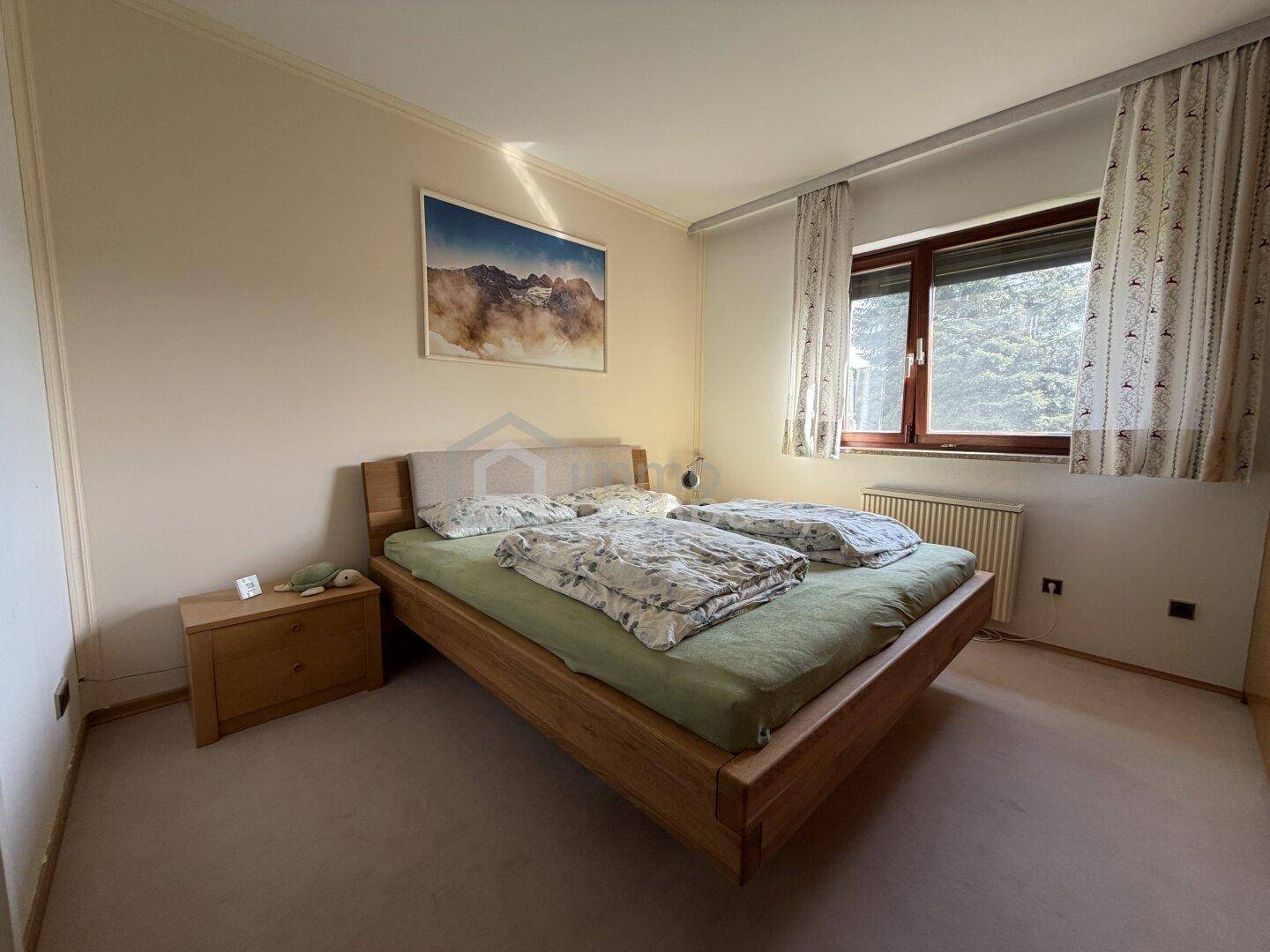 Schlafzimmer mit Holzbett, Teppichboden und Fenster mit Blick auf Bäume, in gutem Zustand.