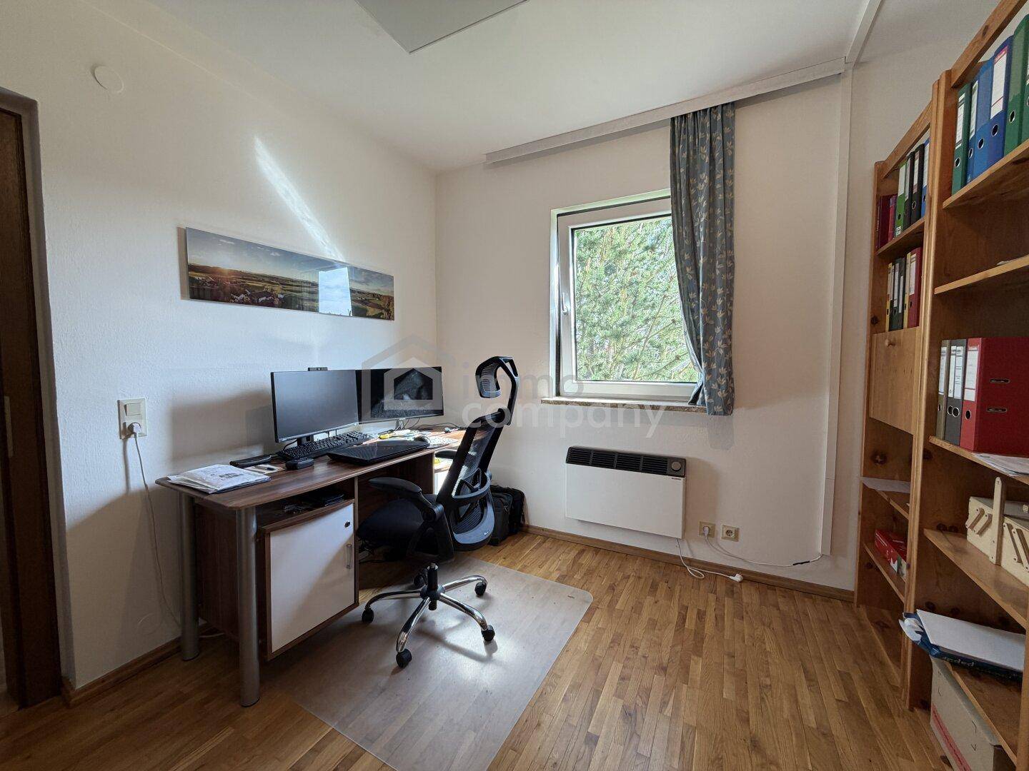 Arbeitszimmer mit Holzparkett, Schreibtisch und Bücherregal, hell durch das Fenster mit Grünblick.