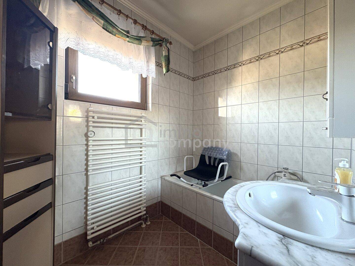 Gefliestes Badezimmer mit Badewanne, Waschbecken und beheiztem Handtuchhalter am Fenster.