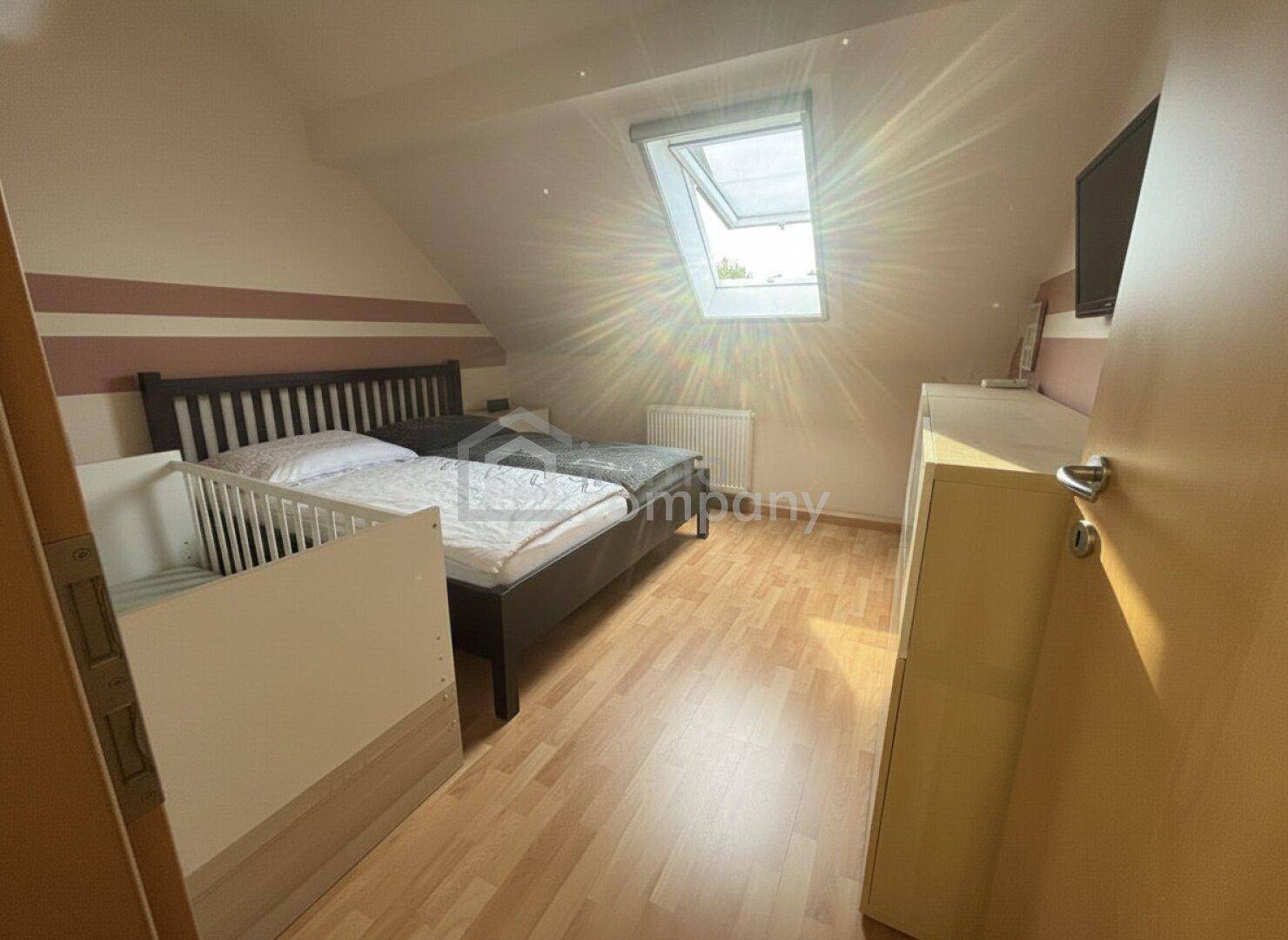Helles Schlafzimmer mit Bett, Babybett, Holzboden und Dachfenster, durchflutet von Sonnenlicht.