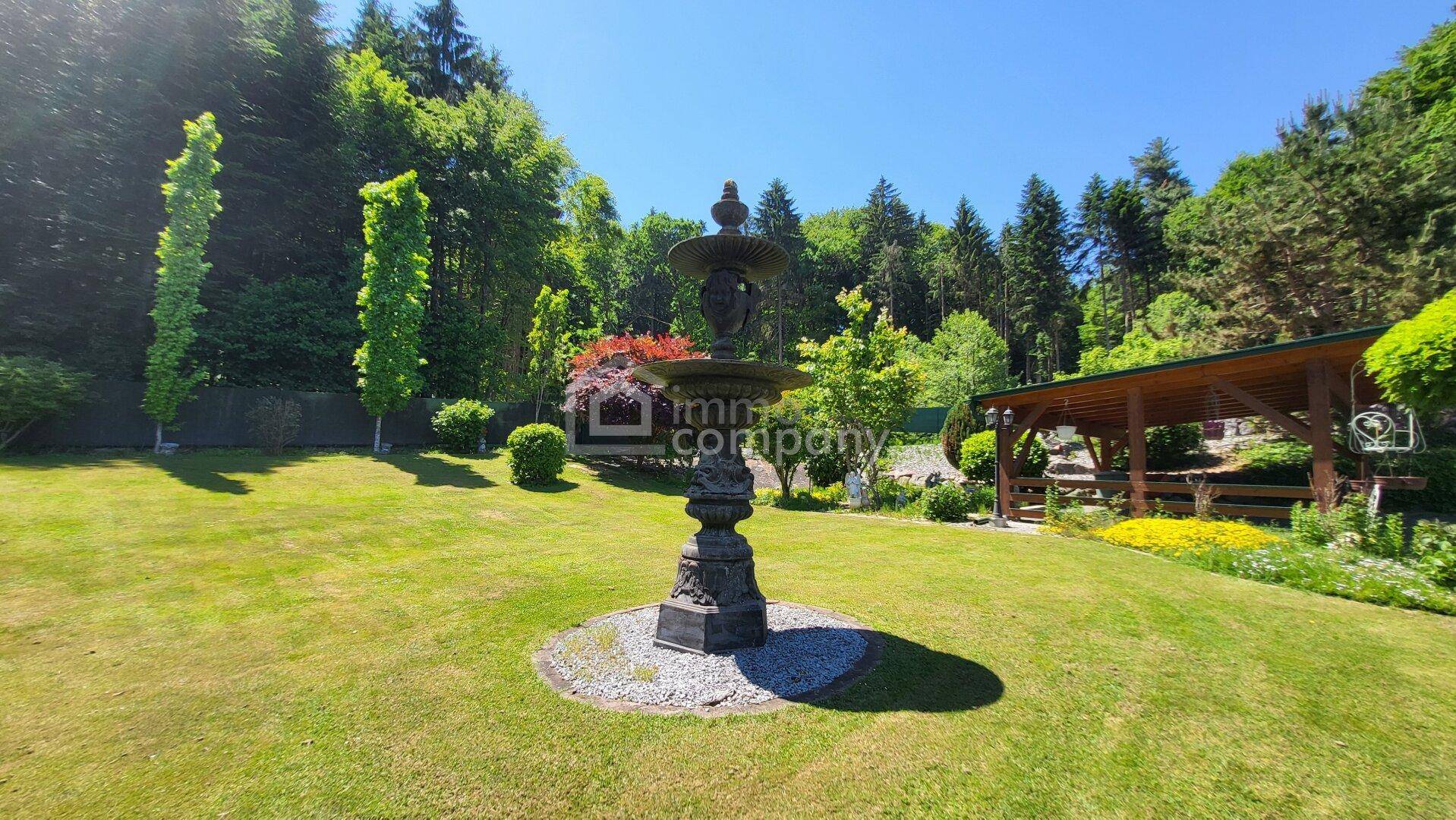Ein dekorativer Brunnen steht auf einer Rasenfläche, umgeben von Bäumen und Sträuchern in einem weitläufigen Garten.