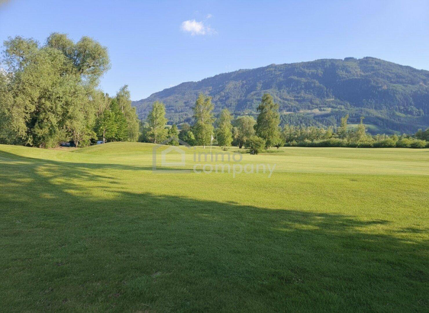 Idyllische Landschaft eines Golfplatzes mit Bäumen, einem Fahnenstock und Bergen im Hintergrund.