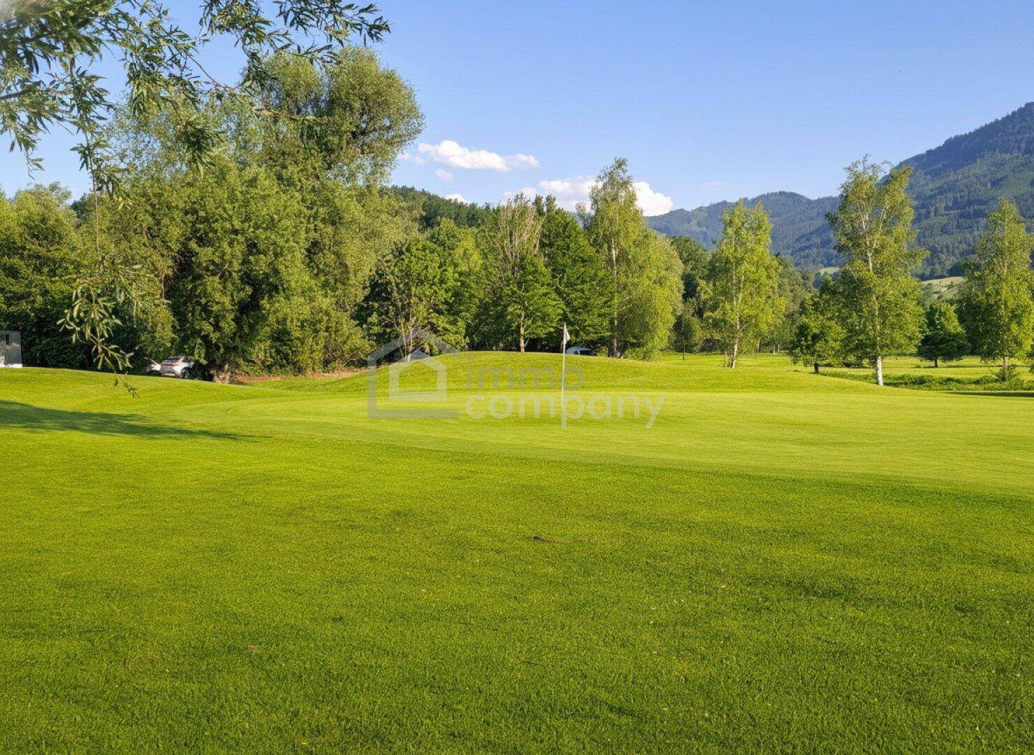 Sonniger Golfplatz mit üppigem Grün, umgeben von Bäumen und einer Bergkulisse.