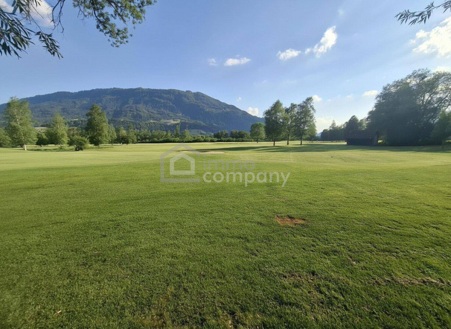 Panoramablick über einen gepflegten Golfplatz mit sanften Hügeln und Bergen am Horizont.