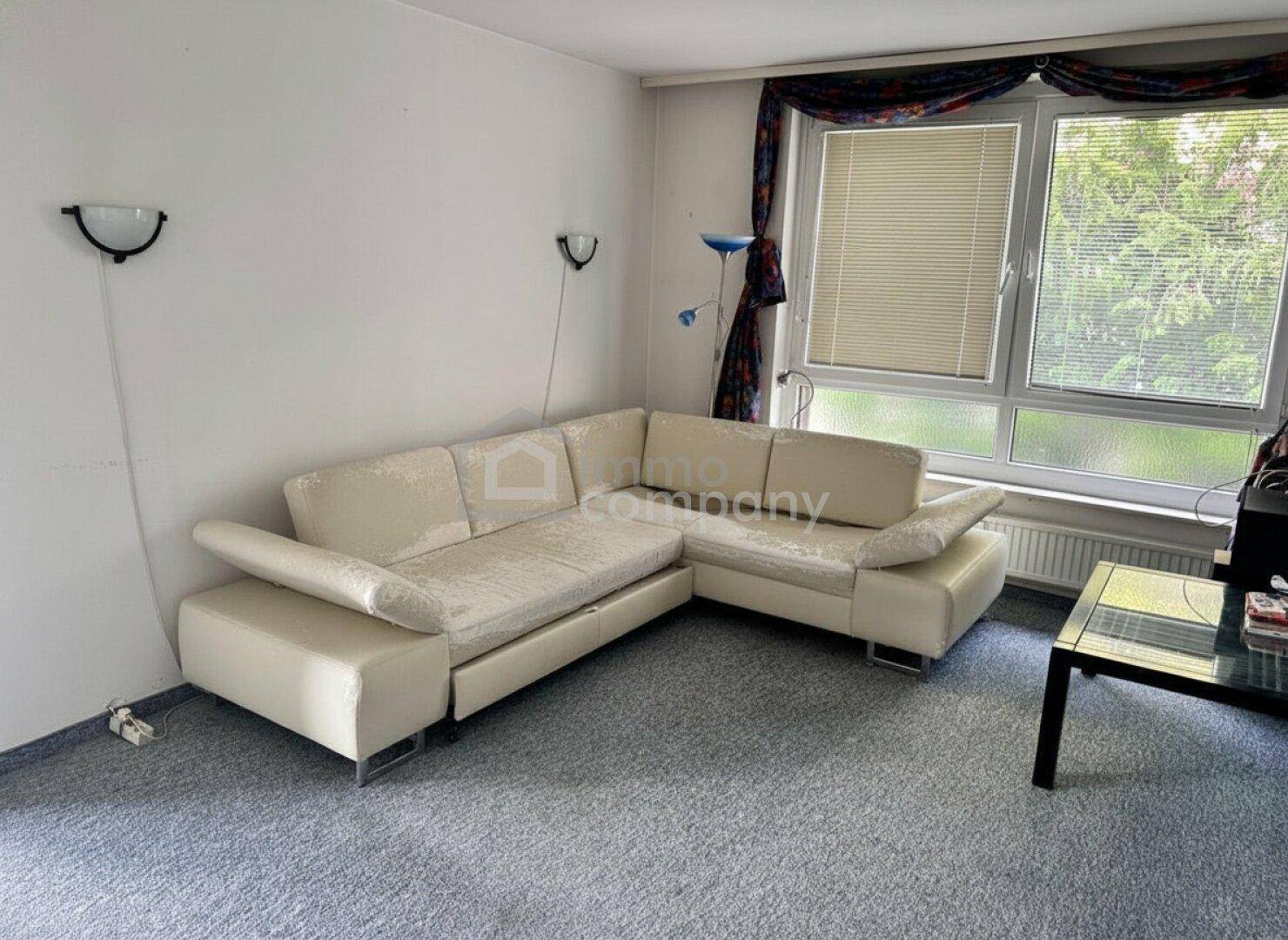 Gemütliches Wohnzimmer mit beigem Ecksofa, Teppichboden und Fenster mit Jalousien und Vorhängen.