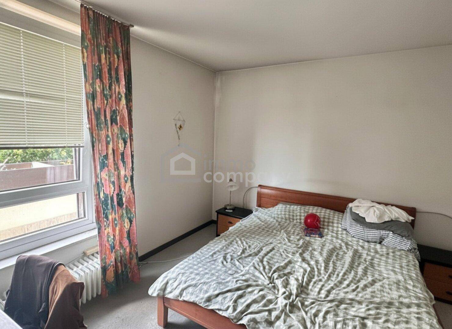 Schlafzimmer mit Holzbett, Nachttischen und einem Fenster mit bunten Vorhängen und Jalousien.