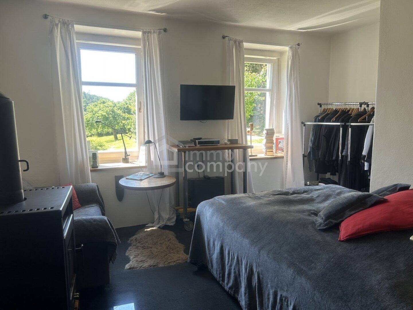 Schlafzimmer mit Bett, Kleiderstange und zwei Fenstern mit Blick ins Grüne.