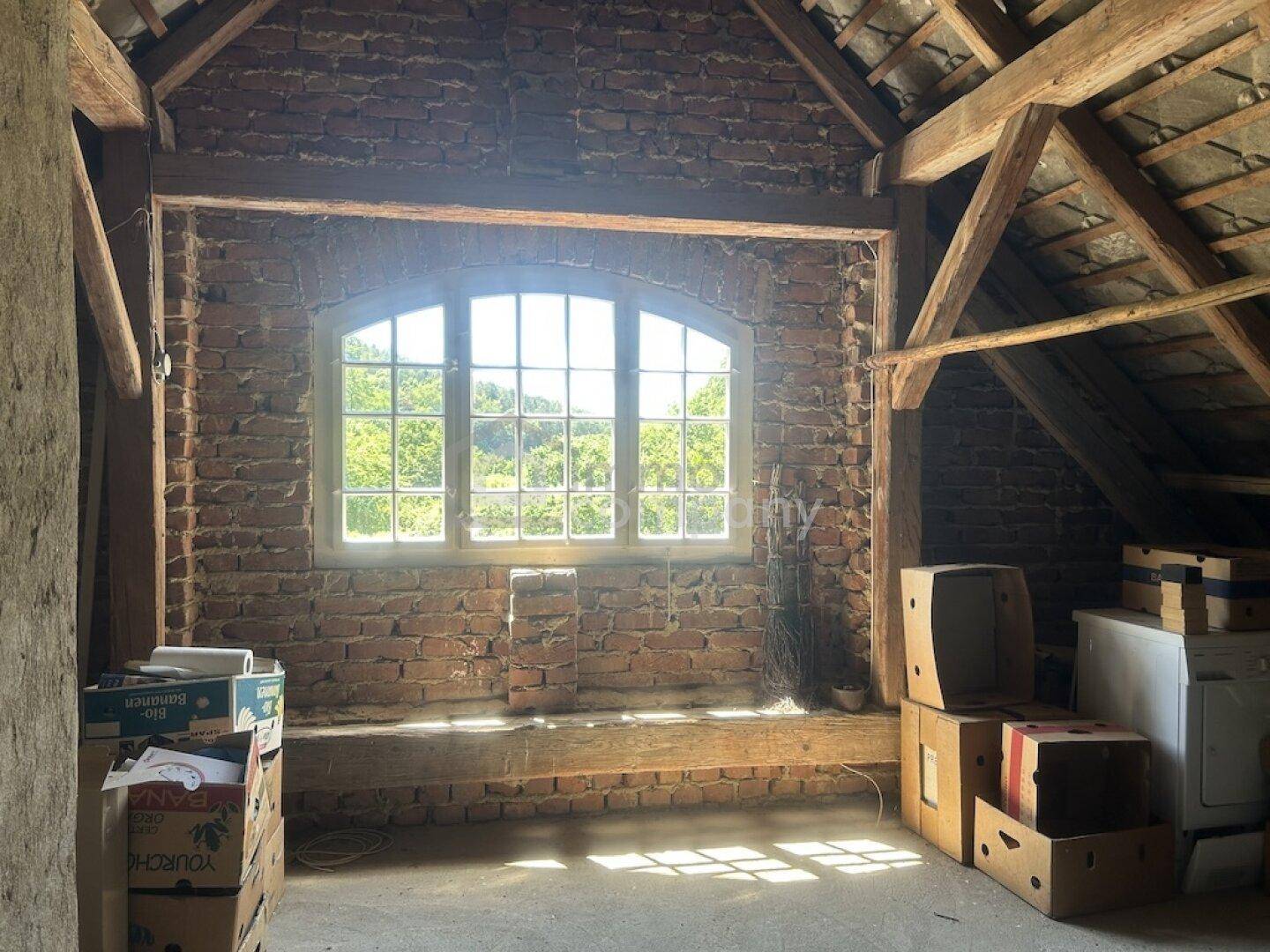 Heller Dachboden mit Ziegelwänden, Holzbalken und einem Fenster mit Blick ins Grüne.