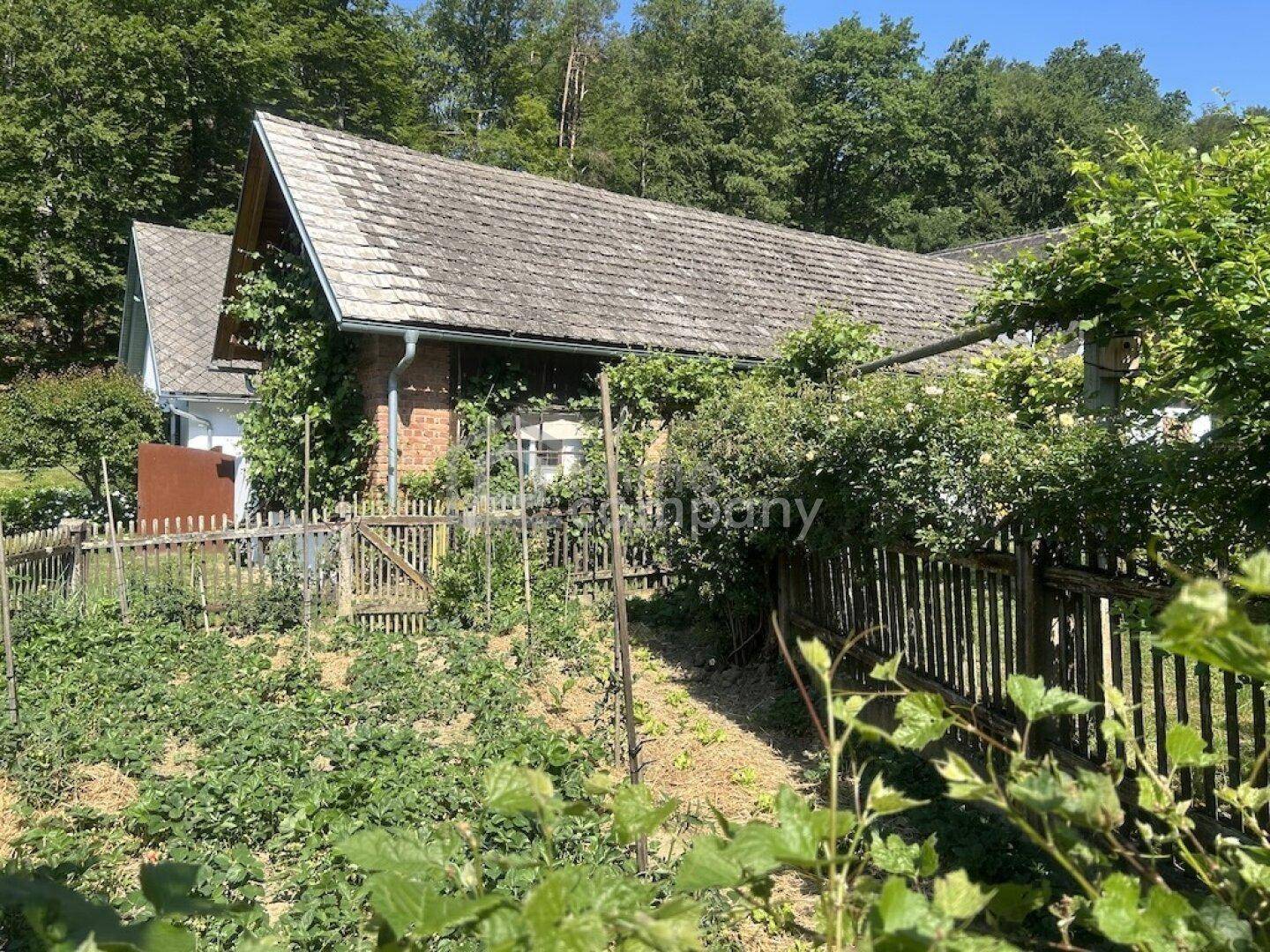 Ländliches Haus mit Satteldach, umgeben von einem Garten mit Zaun und üppiger Vegetation.