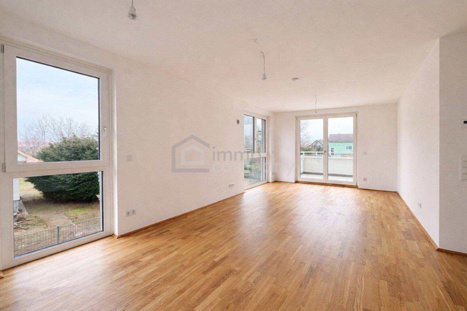 Heller, leerer Wohnbereich mit großen Fenstern, Holzboden und Zugang zu einem Balkon.