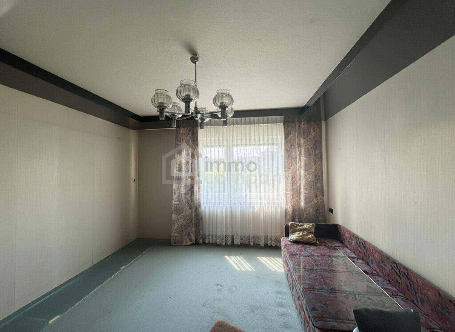 Wohnzimmer mit Teppichboden, langer Couch und Fenster mit gemusterten Vorhängen.