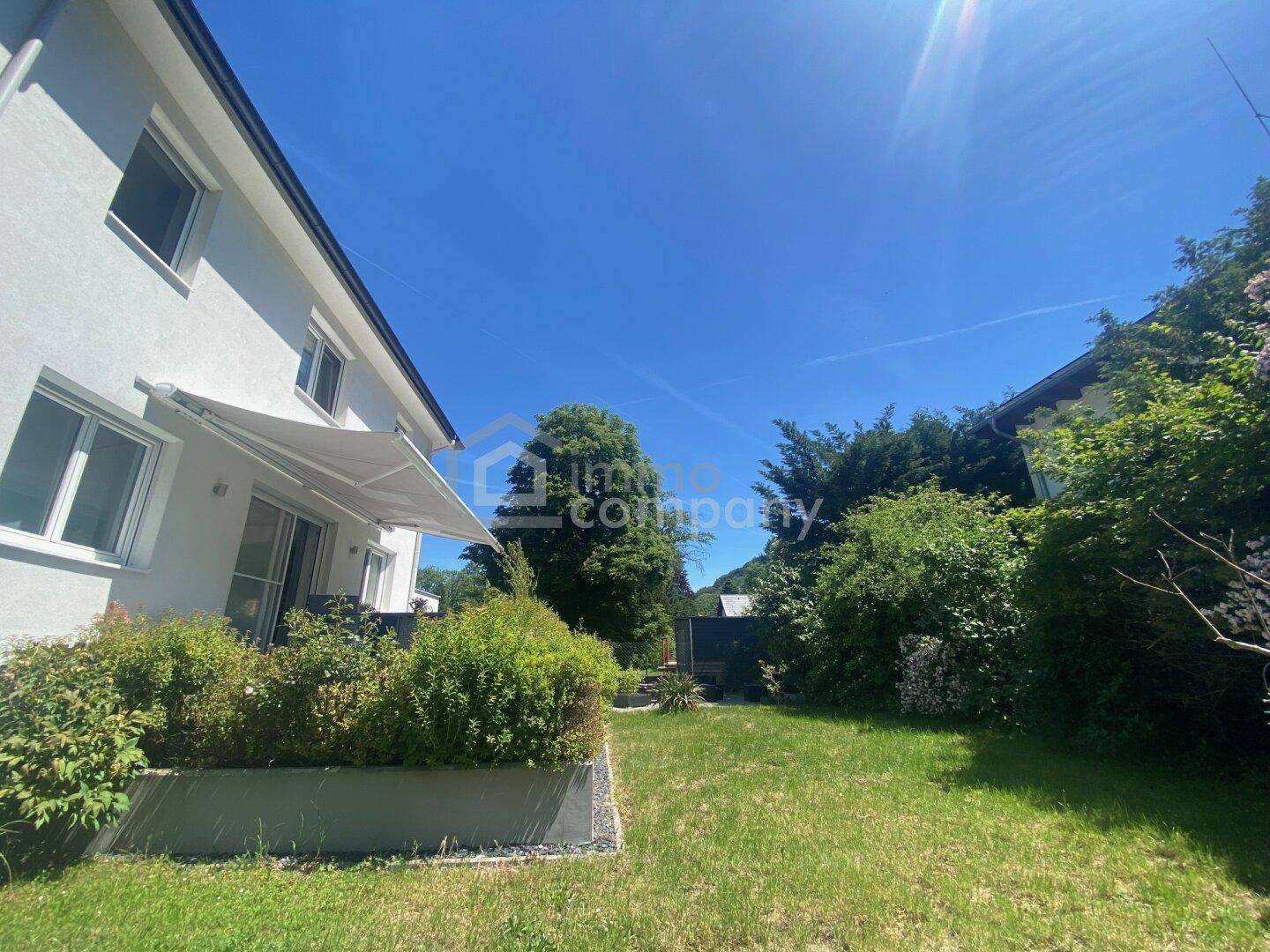 Sonnige Rasenfläche neben dem Haus mit einer ausfahrbaren Markise über der Terrasse und üppigem Grün.