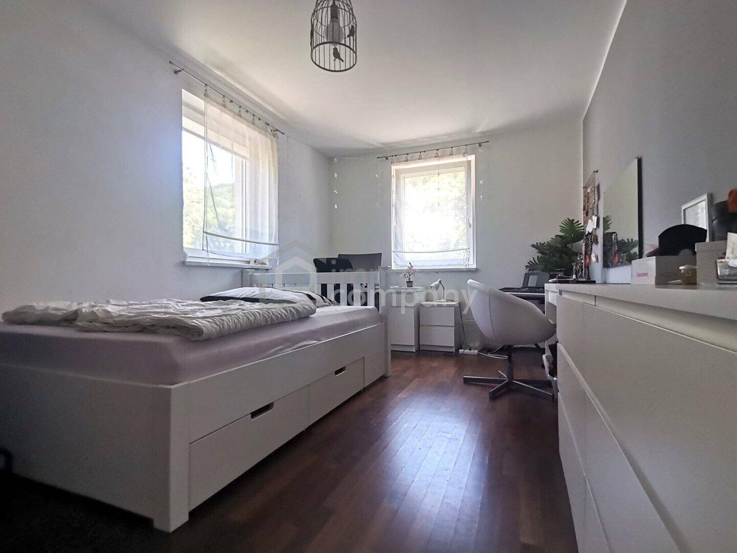 Helles Schlafzimmer mit Holzboden, weißem Bett und Kommode, sowie zwei Fenstern für Tageslicht.