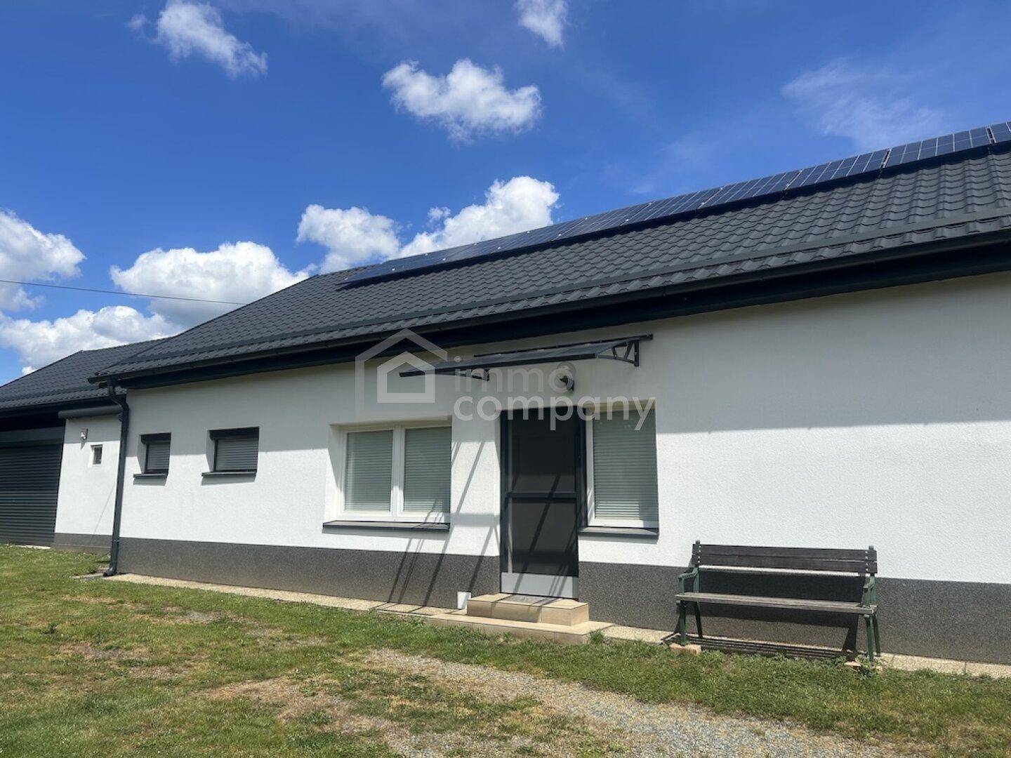 Seitenansicht des Hauses mit Solarpaneelen auf dem Dach und einem kleinen Sitzbereich vor dem Eingang.