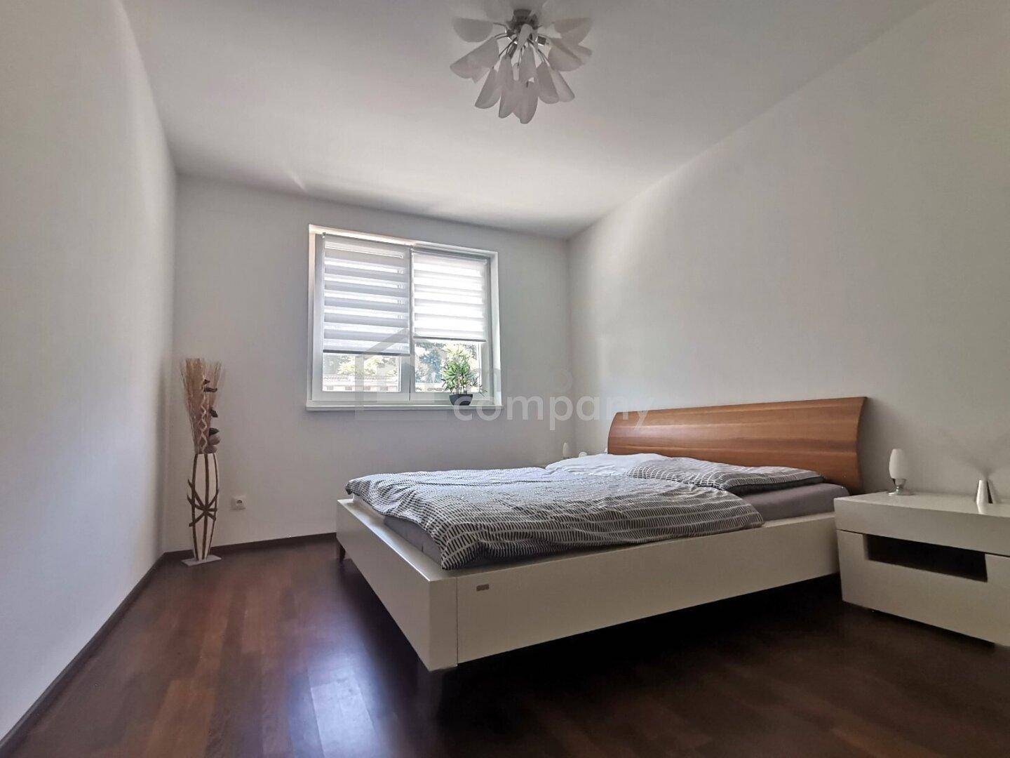 Ruhiges Schlafzimmer mit Holzboden, modernem Bett und Nachttisch, beleuchtet durch ein großes Fenster.