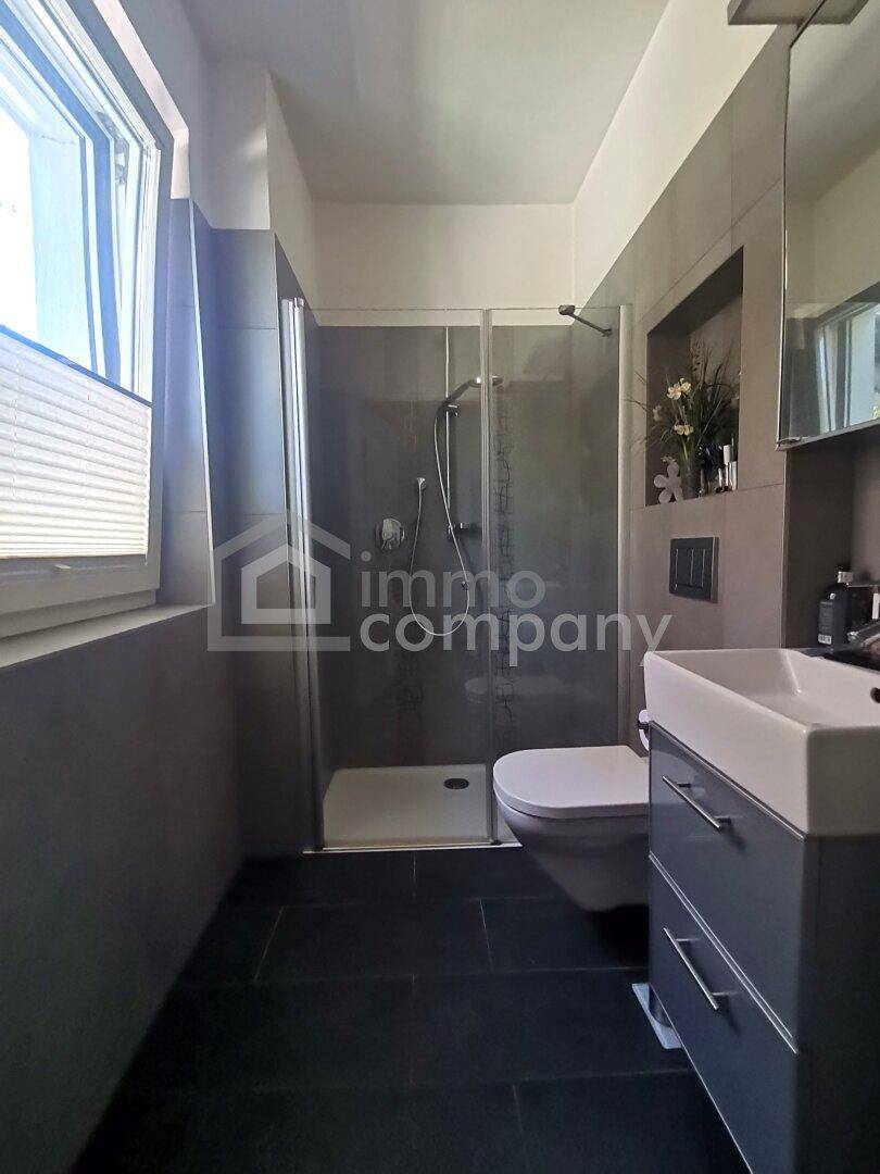 Modernes Badezimmer mit Duschkabine, wandhängendem WC und Fenster für natürliche Belüftung.
