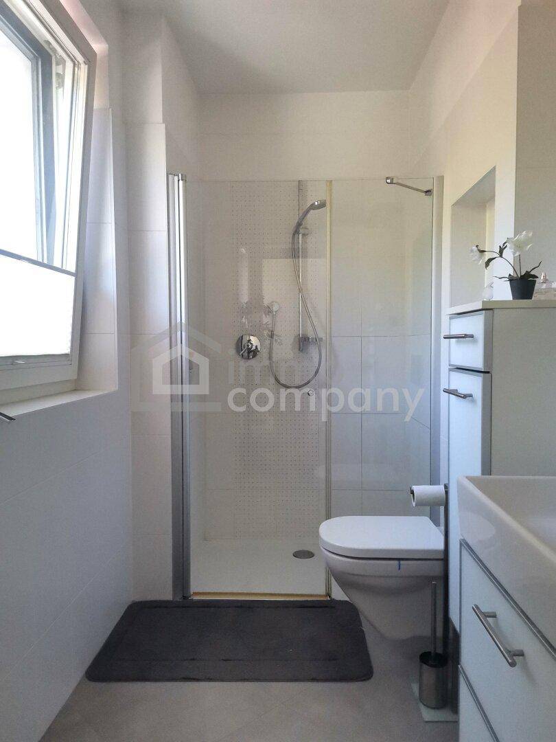 Helles Badezimmer mit Duschkabine, WC und Fenster, ausgestattet mit weißen Fliesen und modernem Design.