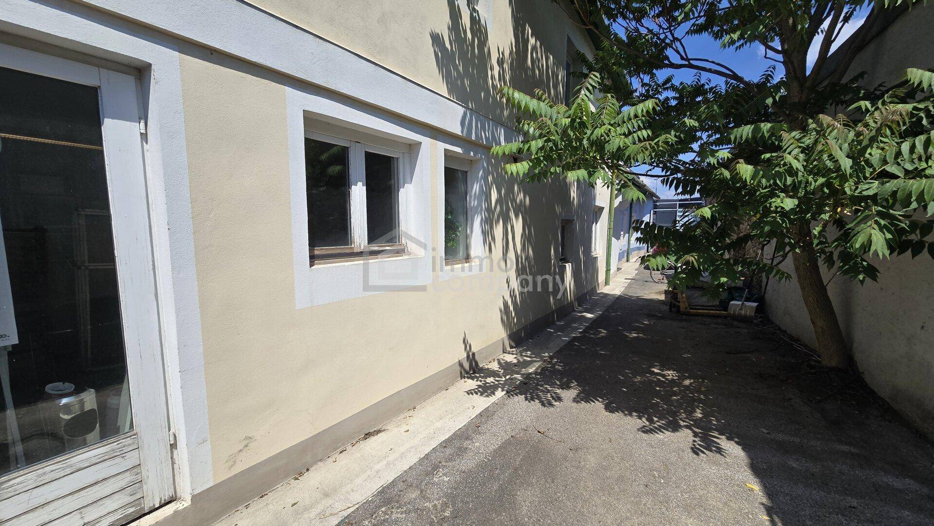 Seitenansicht eines Gebäudes mit beiger Fassade und Fenstern, entlang eines schmalen Weges mit Schattenwurf von Bäumen.