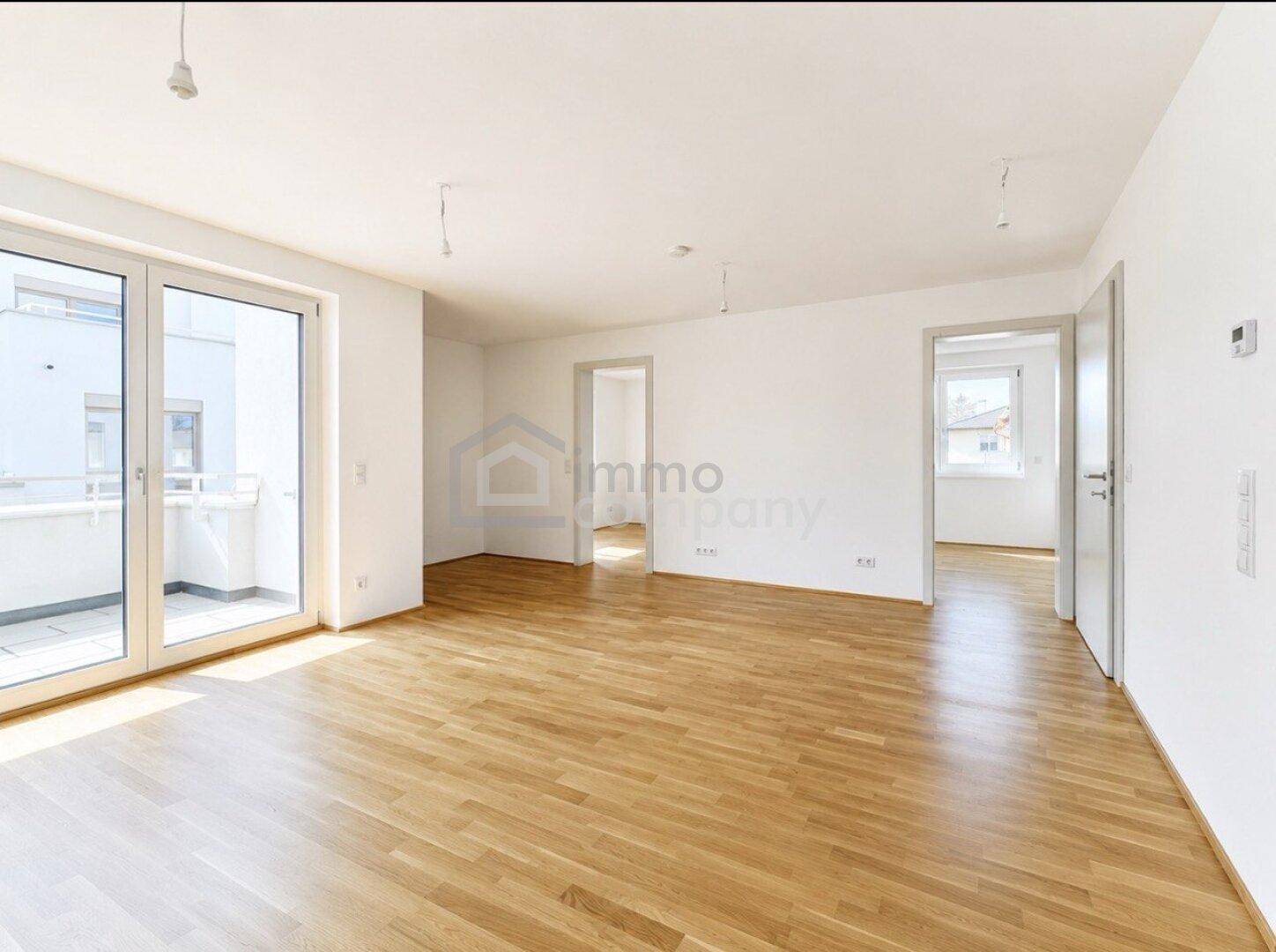 Geräumiger, leerer Wohnbereich mit hellem Parkettboden und großen Fenstern zum Balkon.