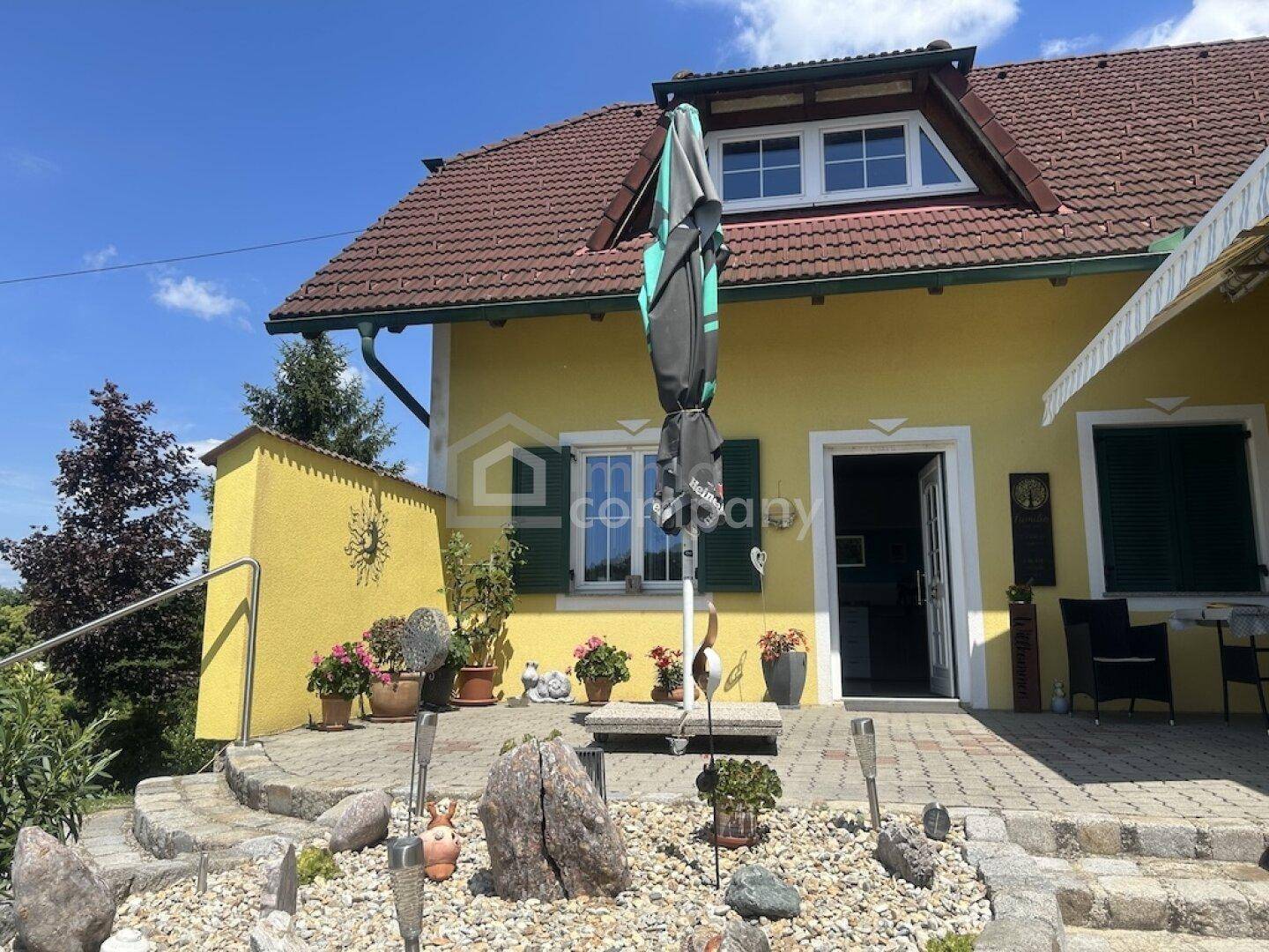 Sonniger Terrassenbereich vor dem Haus mit dekorativen Steinen, Blumen und einem großen Sonnenschirm.