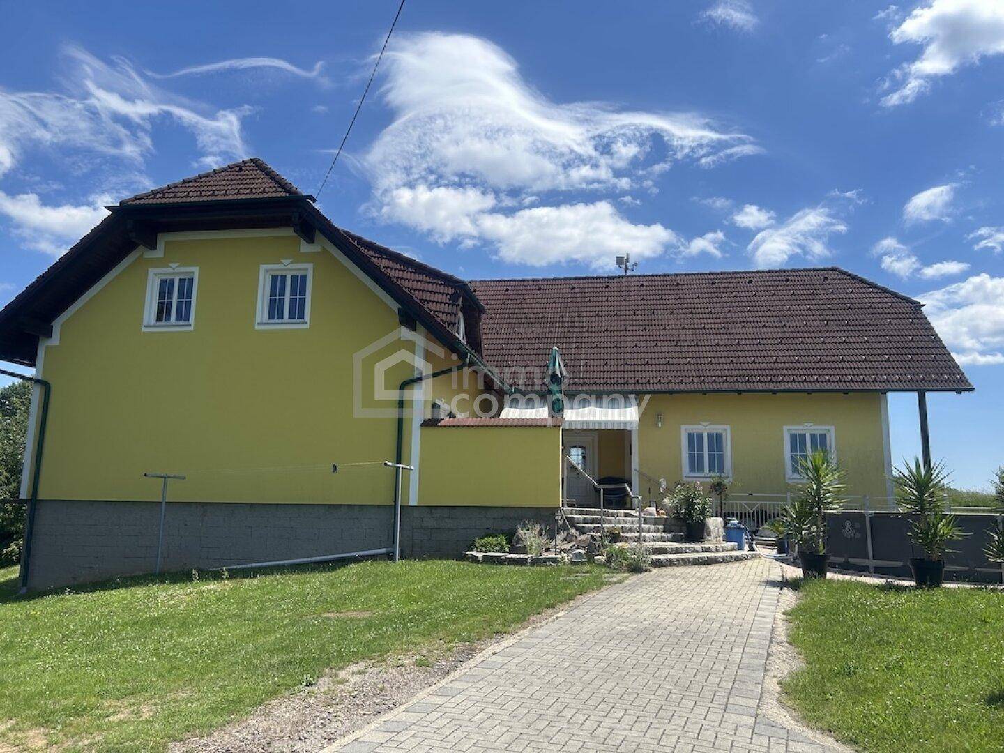 Ansicht des gelben Hauses mit braunem Dach, gepflasterter Auffahrt und grüner Rasenfläche.