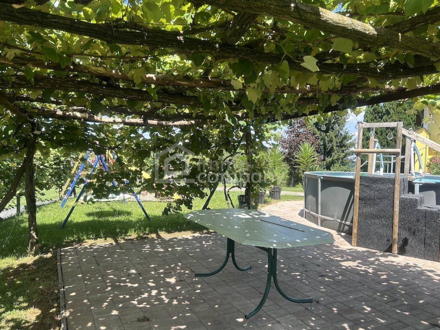 Überdachter Sitzbereich im Garten unter einer grünen Pergola mit Blick auf den Poolbereich.