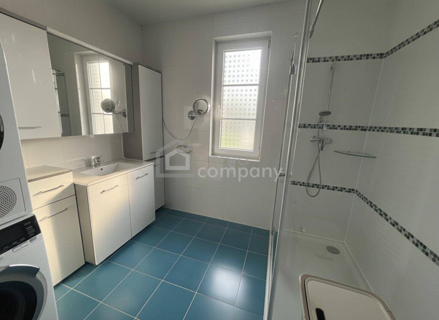 Helles Badezimmer mit blauen Bodenfliesen, Dusche, Waschbecken und Fenster.