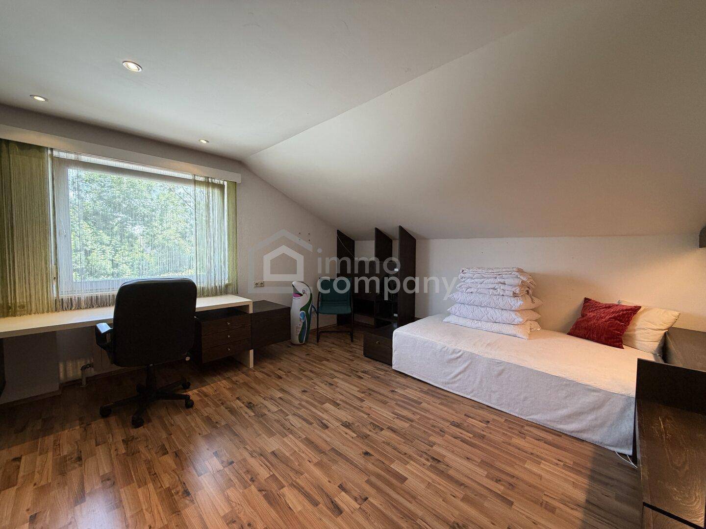 Helles Schlafzimmer im Dachgeschoss mit Holzboden, großem Fenster und Arbeitsbereich.