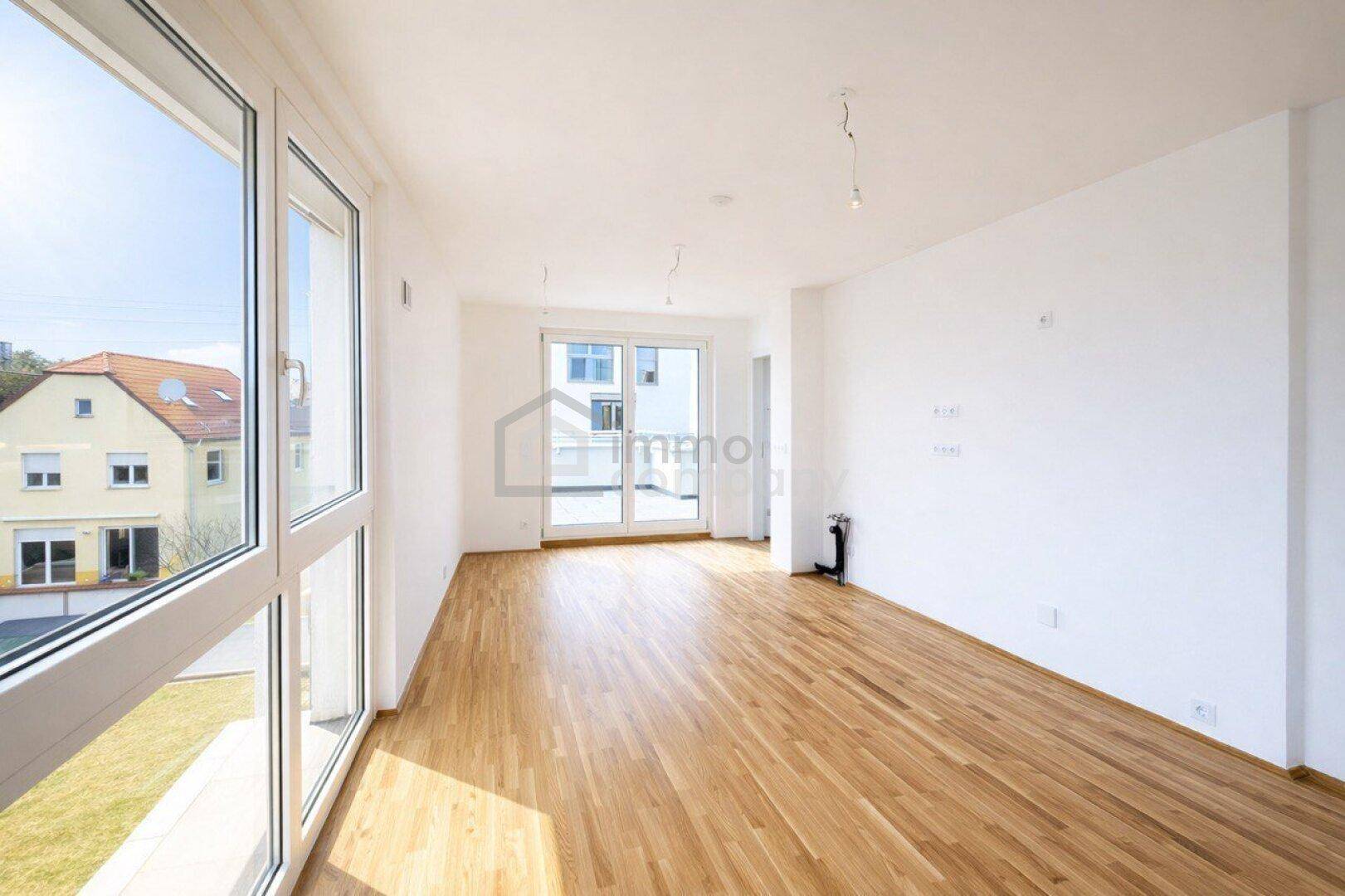 Heller, leerer Wohnbereich mit Holzboden und großen Fenstern, die viel Tageslicht hereinlassen.