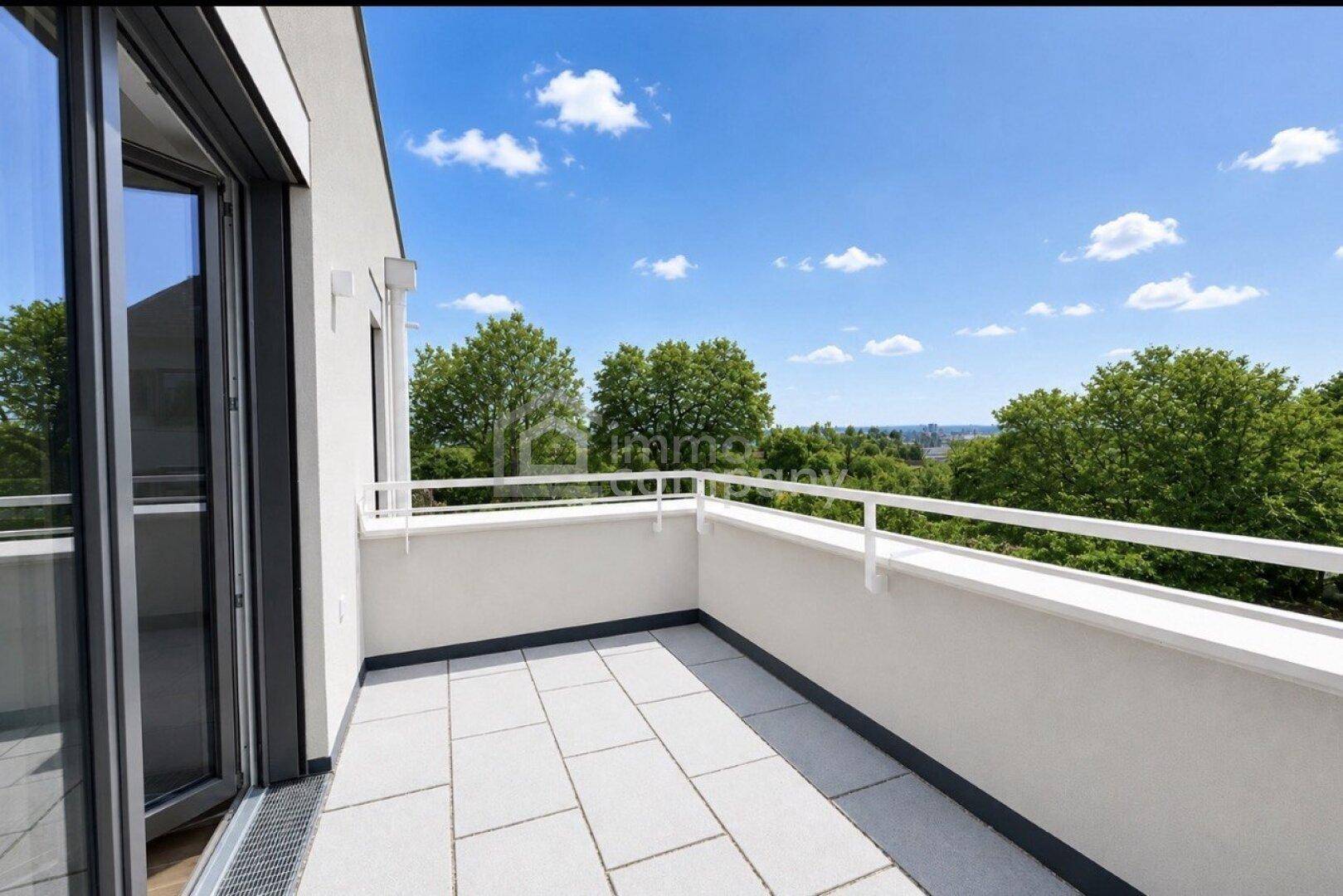 Geräumiger Balkon mit hellen Fliesen, weißem Geländer und weitem Blick ins Grüne unter blauem Himmel.