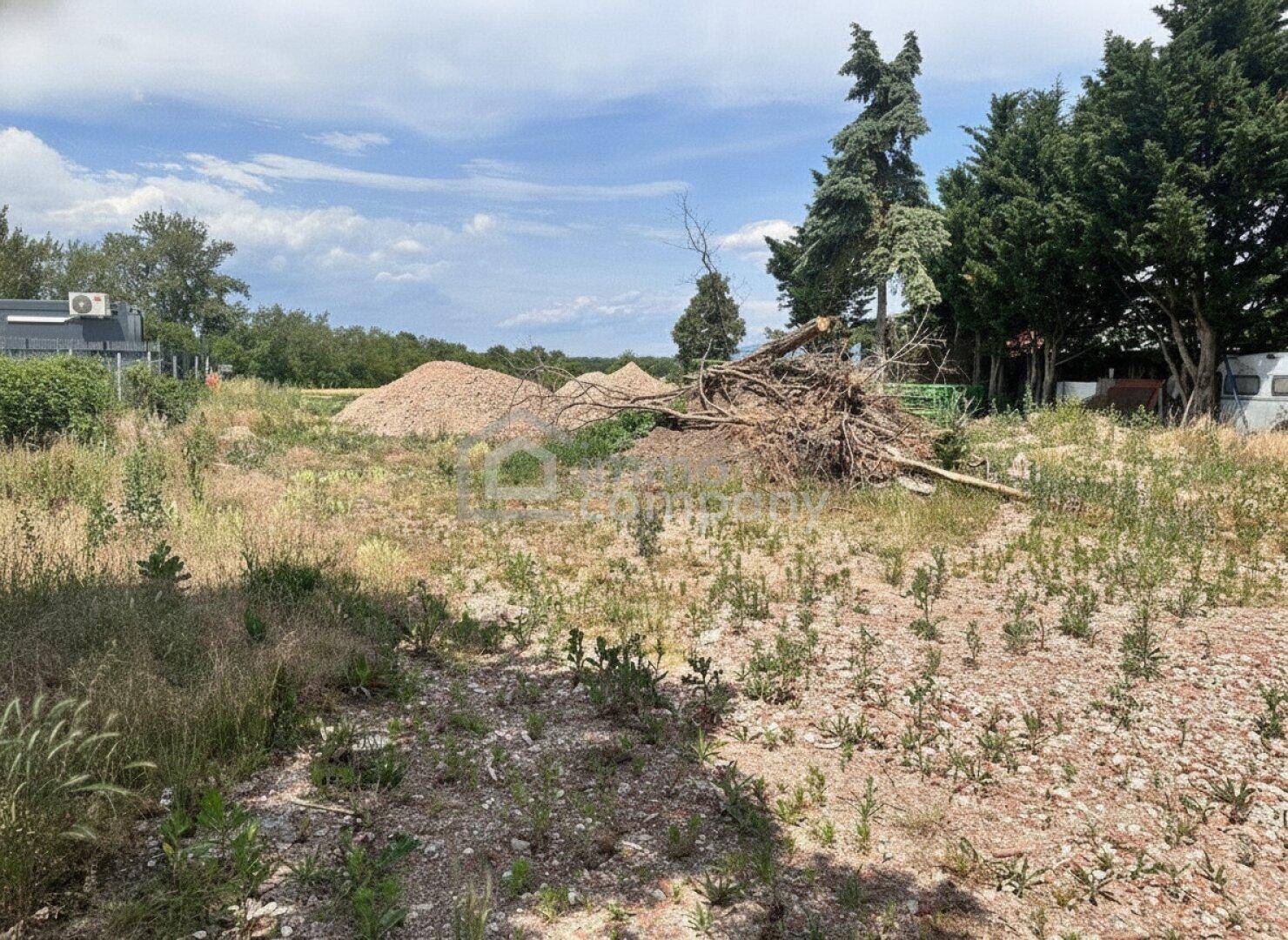 Unbebautes Grundstück mit Schotter, Erdhaufen und Bäumen im Hintergrund unter bewölktem Himmel.