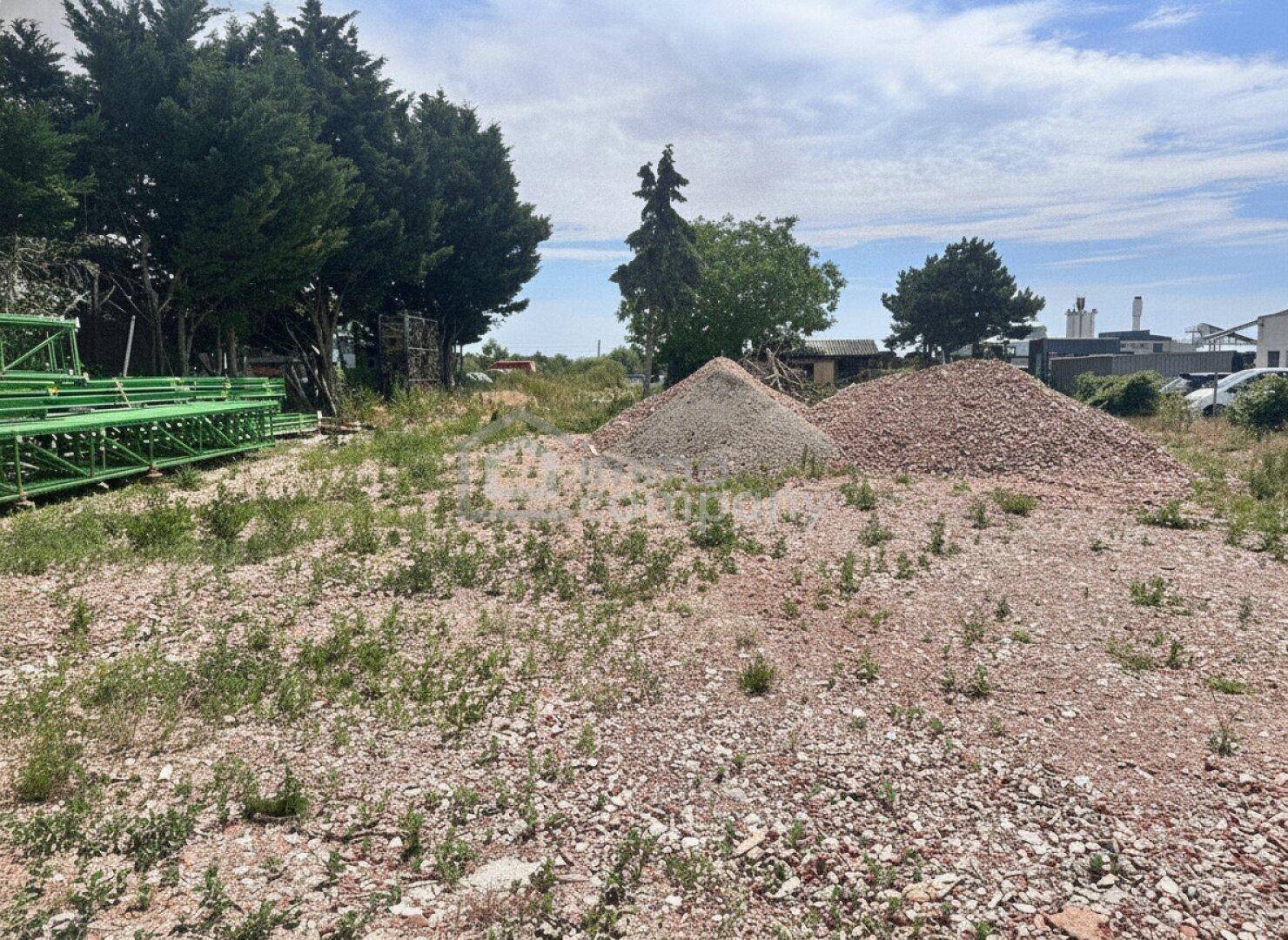 Weitläufiges Grundstück mit Schotter, Erdhaufen und dichter Vegetation unter blauem Himmel.