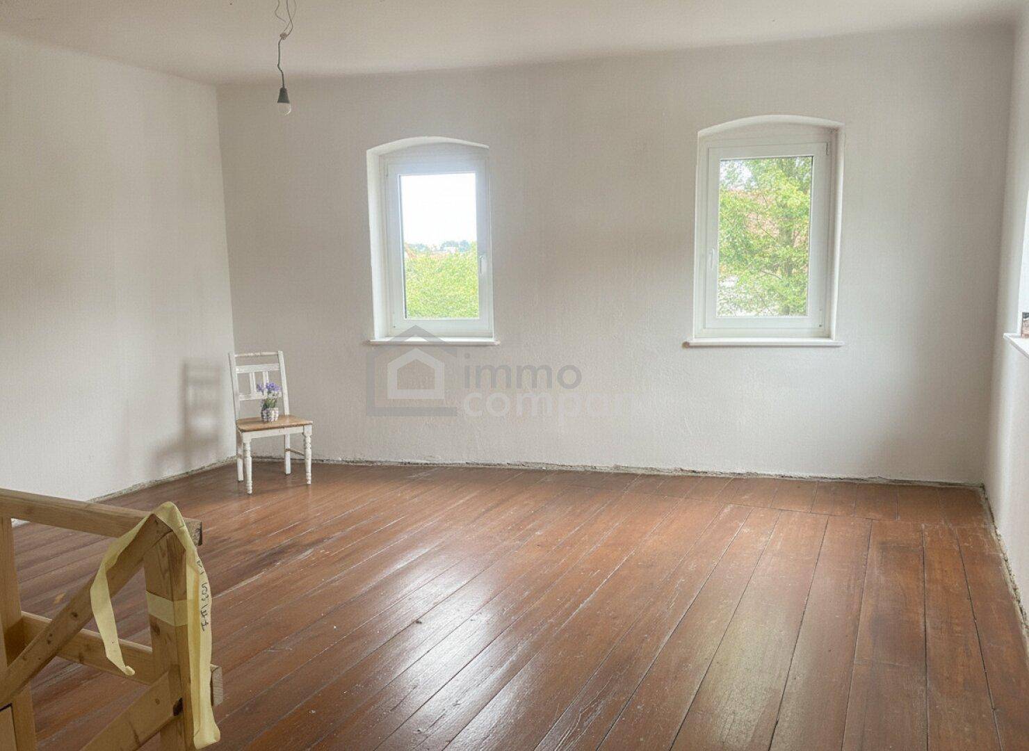 Geräumiges Zimmer mit Holzdielenboden, weißen Wänden und zwei großen Fenstern, viel Tageslicht.