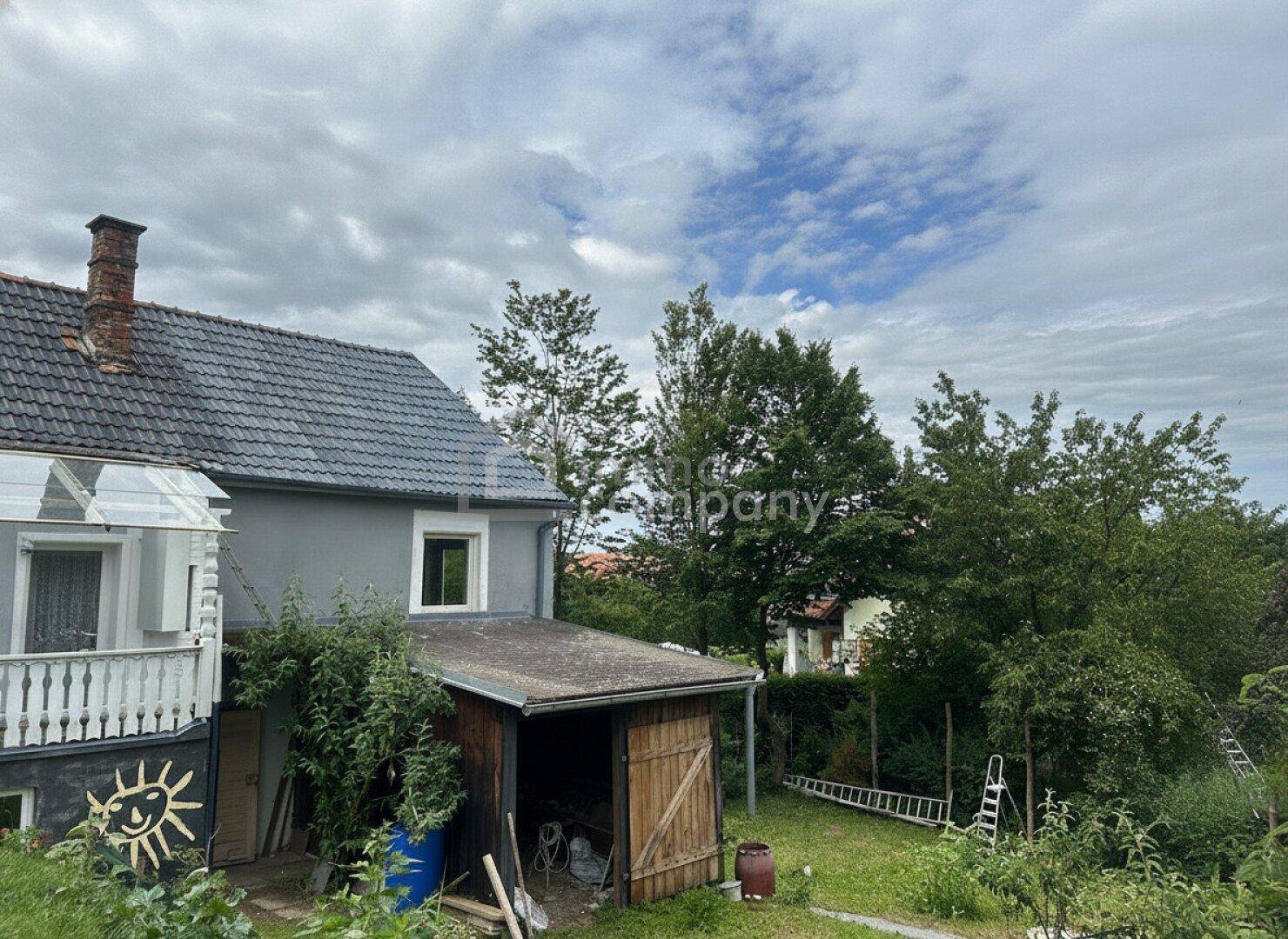Rückansicht eines Hauses mit grauem Putz, einem Holzschuppen und einem grünen Gartenbereich unter bewölktem Himmel.