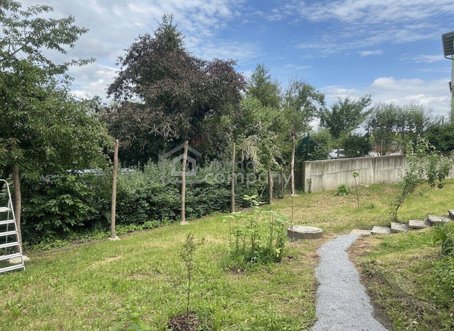 Grüner Gartenbereich mit Rasenfläche, Bäumen und einem Kiesweg, umgeben von Natur und einem Zaun.
