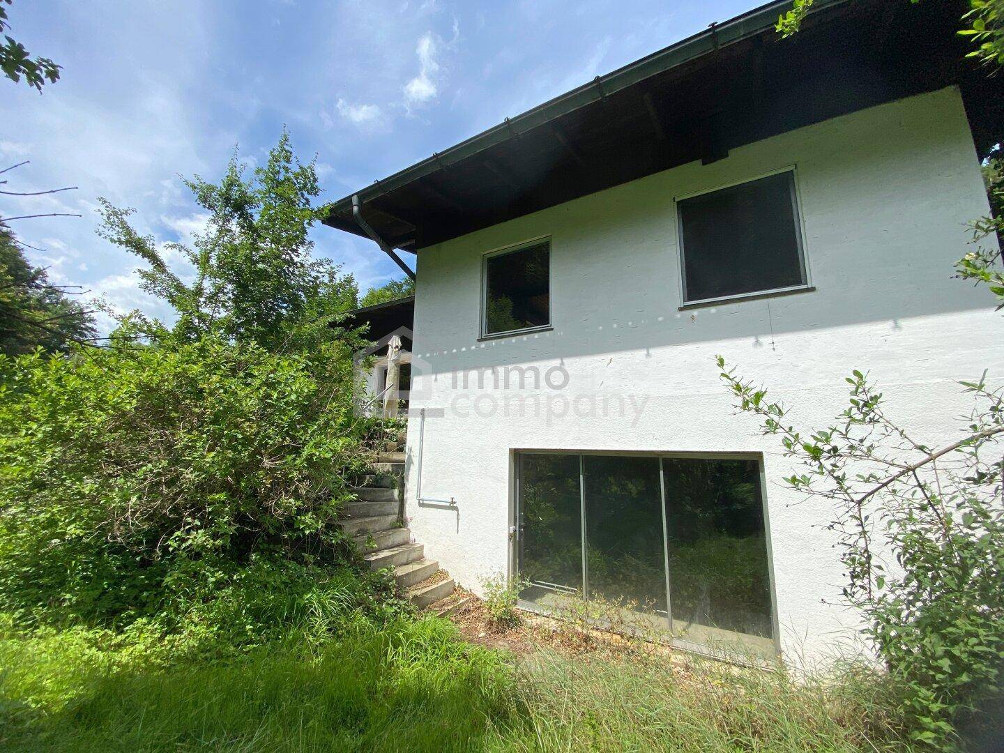 Fassade des Hauses mit Fenstern, Außentreppe und umgeben von üppigem Grün.