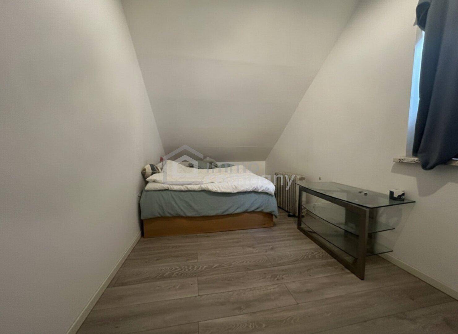 Schlafzimmer mit Bett, Glasregal und Dachschräge, gut beleuchtet durch ein Fenster.