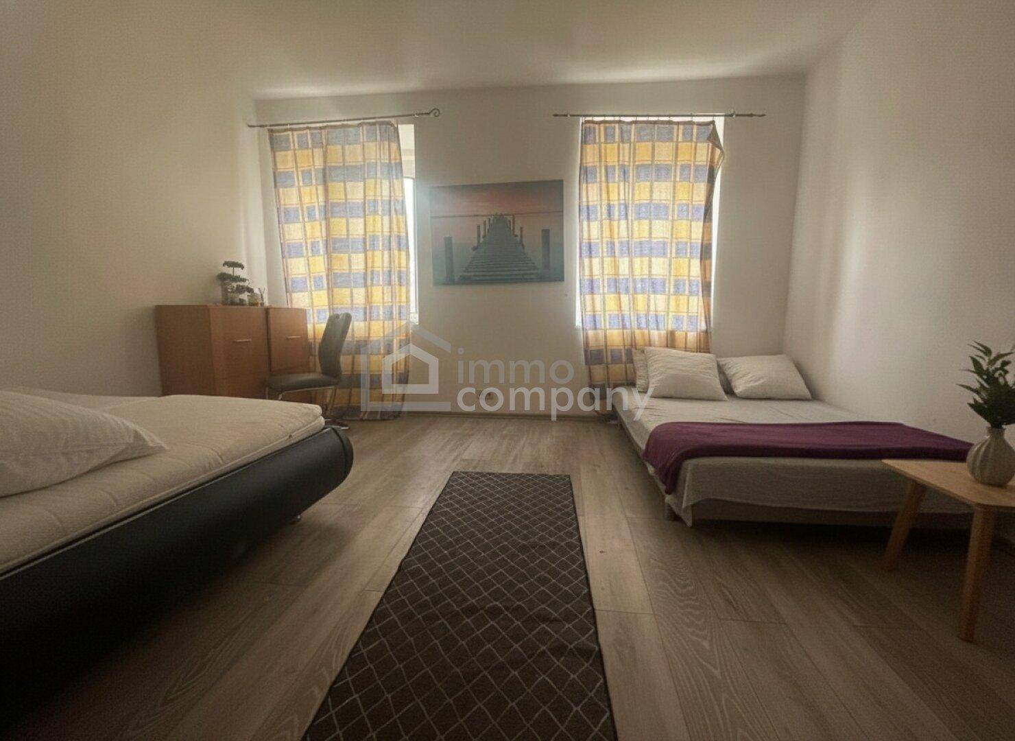 Helles Schlafzimmer mit zwei Betten, Laminatboden und farbenfrohen Vorhängen an den Fenstern.