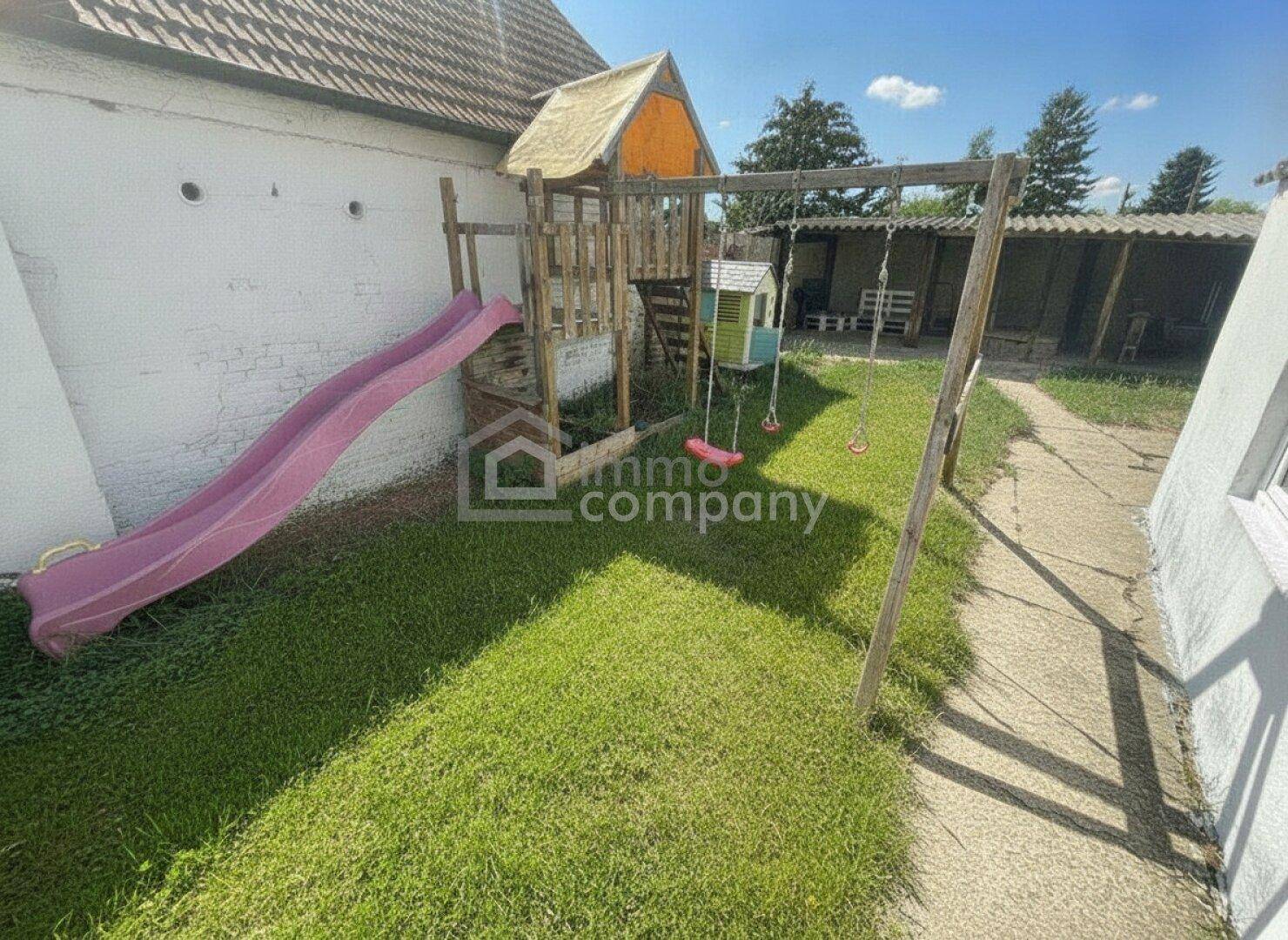Gartenbereich mit Kinderspielplatz, Rutsche und Schaukeln, umgeben von Grünflächen.